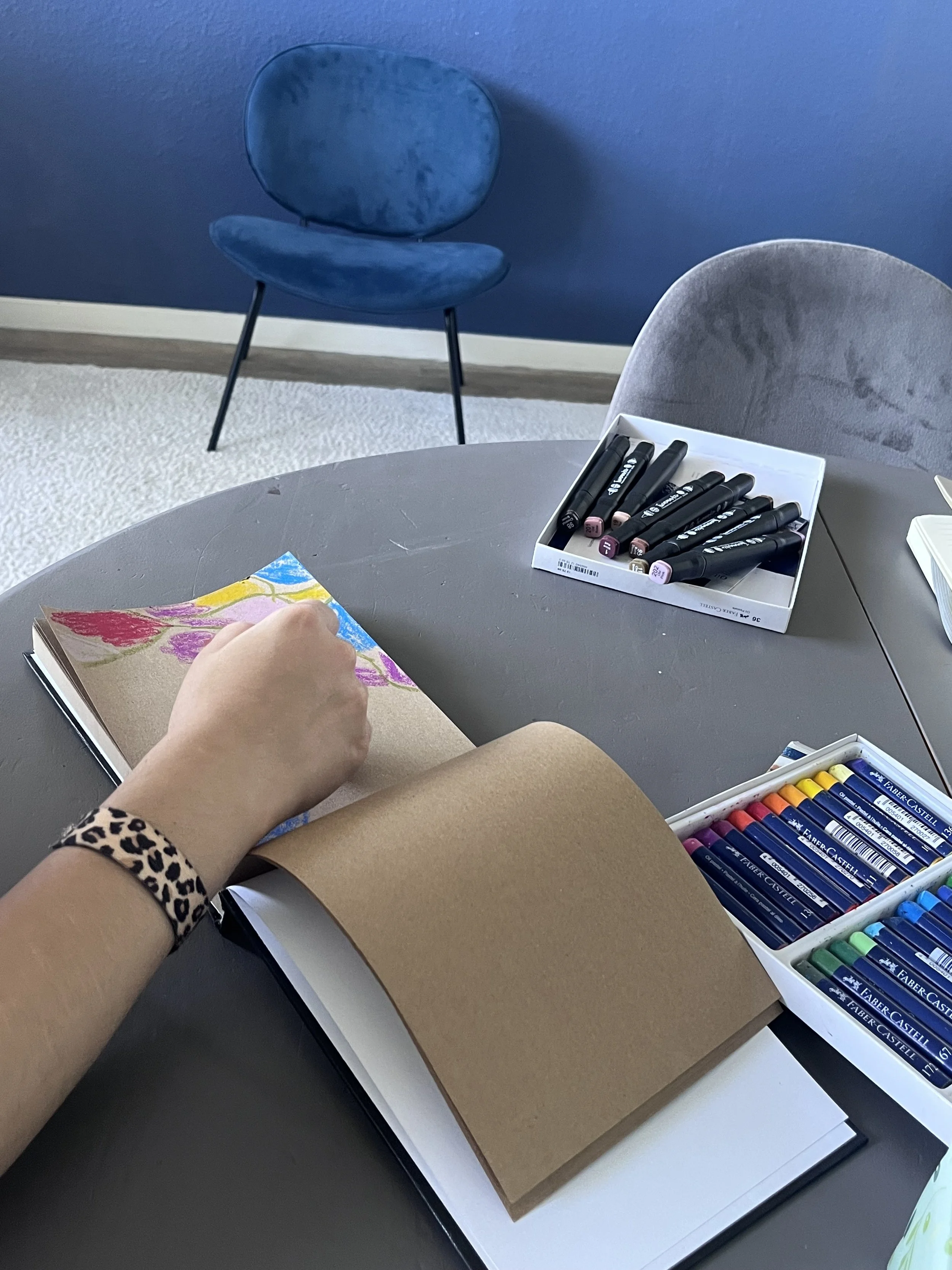 Une personne en train de colorier dans un carnet avec des pastels secs. Sur la table, il y a une boîte de feutres et des crayons de couleur. En arrière-plan, une chaise bleue et une autre chaise grise sont visibles.