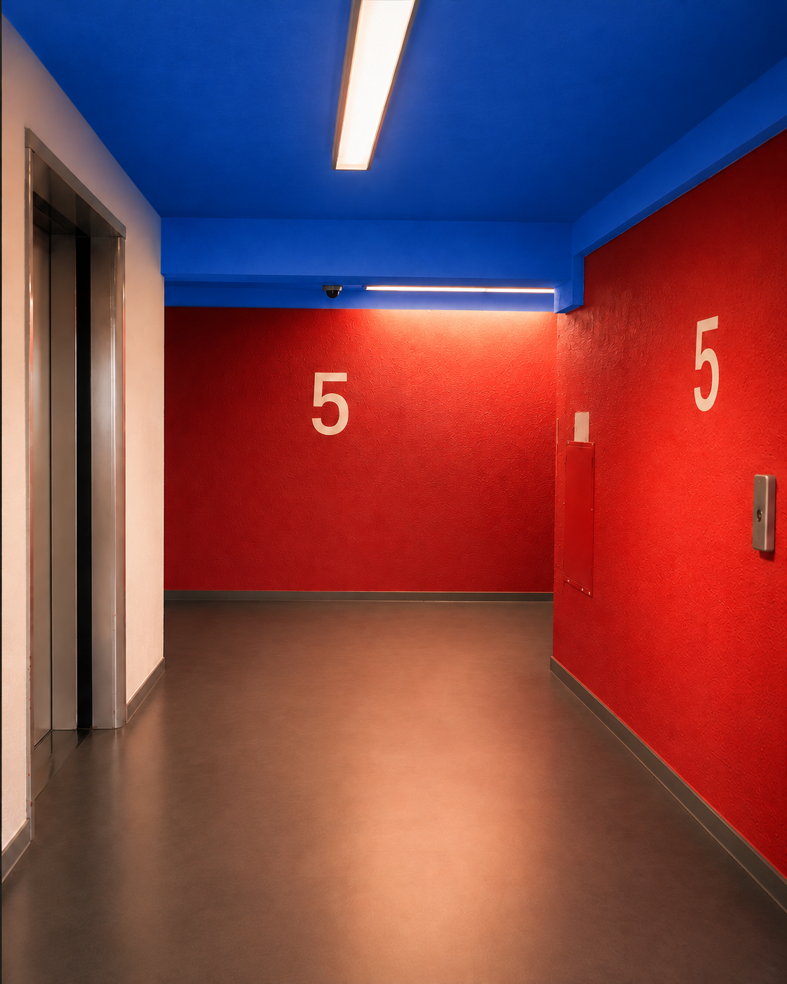 Un couloir d'immeuble avec deux ascenseurs et un mur rouge portant le numéro 5, le plafond est bleu et éclairé par des lumières encastrées indiquent le niveau d'accès.