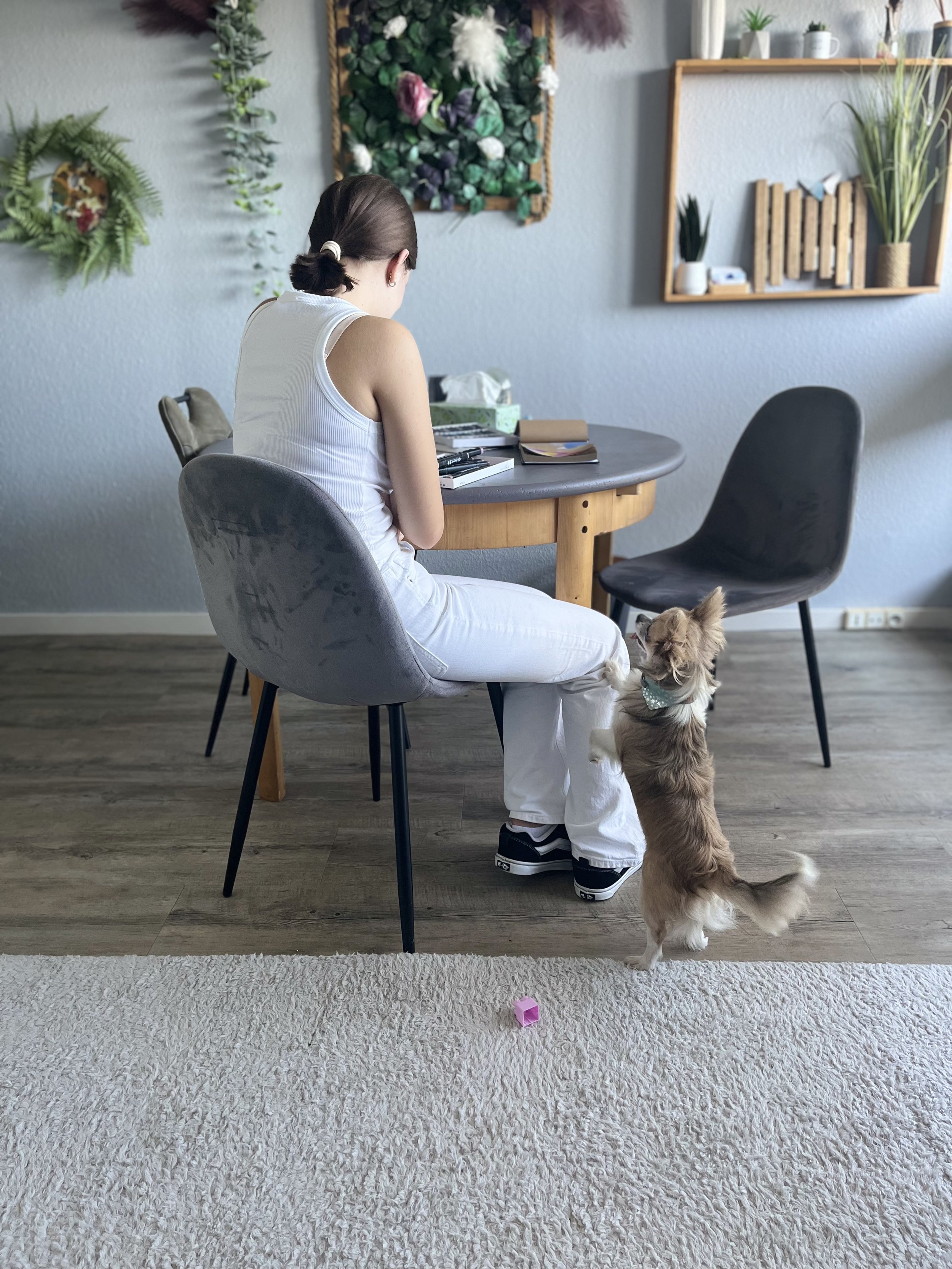 Une jeune femme dessine assise de trois quart à une table en bois dans une pièce décorée avec des plantes et des œuvres d'art murales. Un chien debout sur ses pattes arrière regarde la jeune femme.