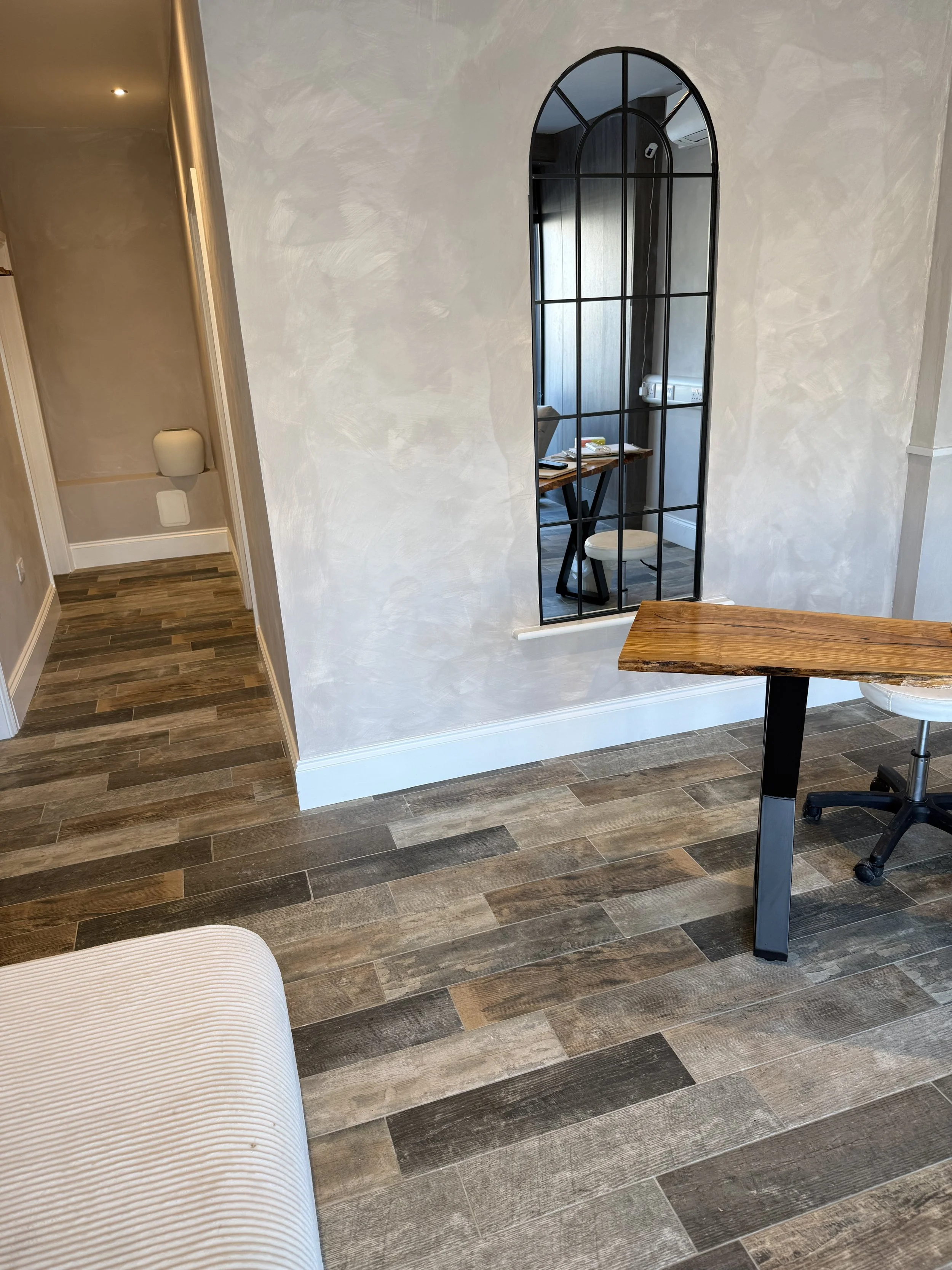 Interior room with wood-look flooring, a white textured wall, a large black-framed arched mirror, a wooden table, a white stool, and part of a beige upholstered chair.
