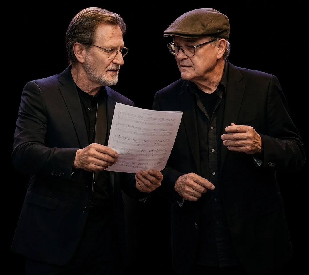 Hank Hehmsoth & John Mills are engaged in a conversation, with one of them holding a sheet of music. They are looking closely at each other against a plain black background.