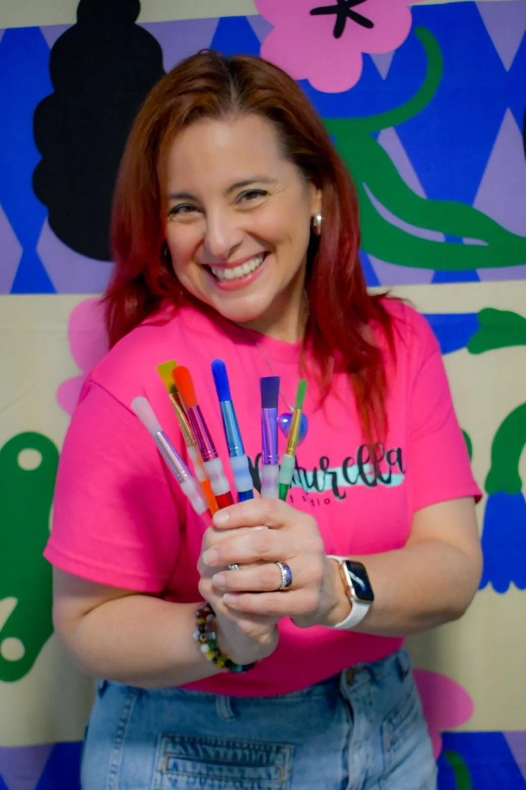 A woman smiling and holding colorful pens in front of a vibrant, colorful background.