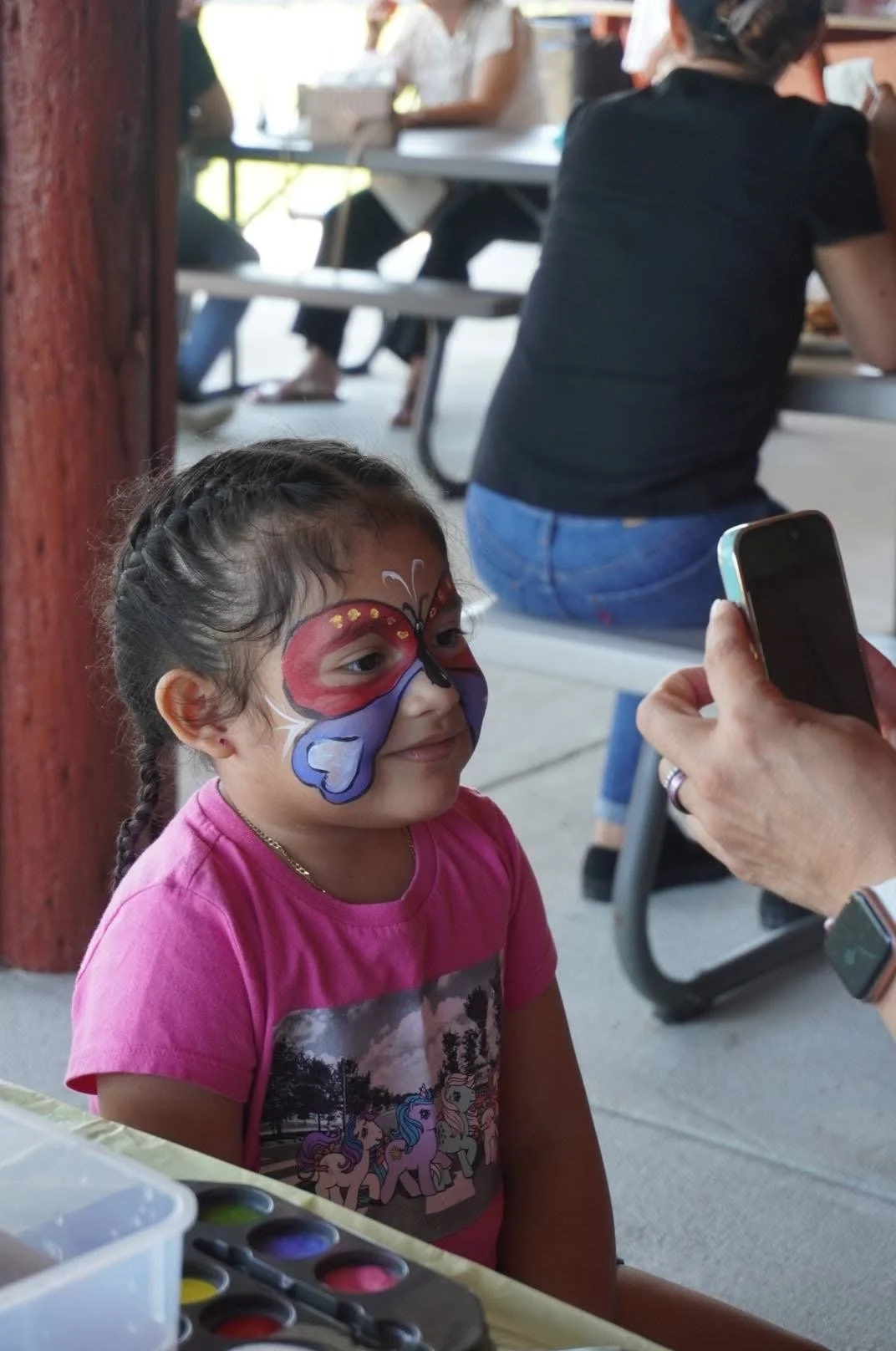 A young girl with face paint of a colorful butterfly is sitting at a table, smiling as someone takes her photo with a smartphone. Other people are seated at picnic tables in the background at an outdoor event.