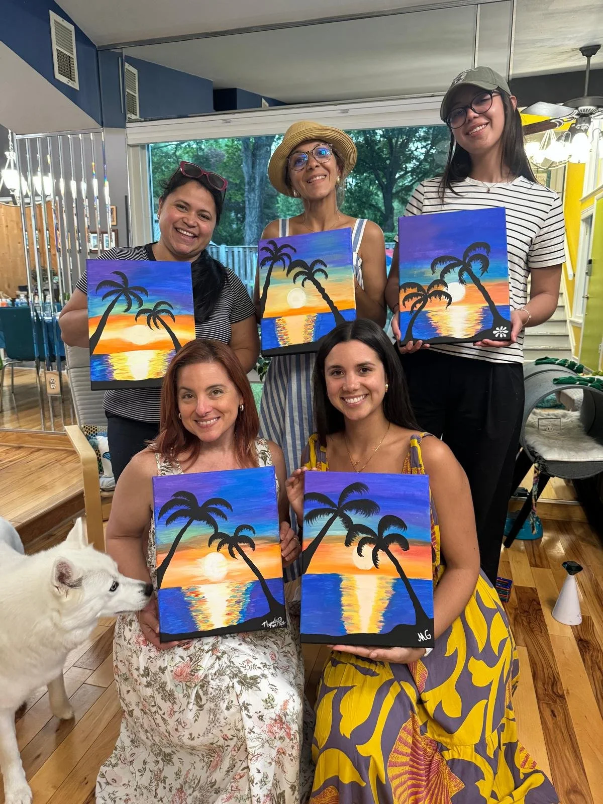 Group of five women and one dog holding artwork of a sunset with palm trees on canvas, inside a brightly lit room with large window, table and chairs, and plants outside.