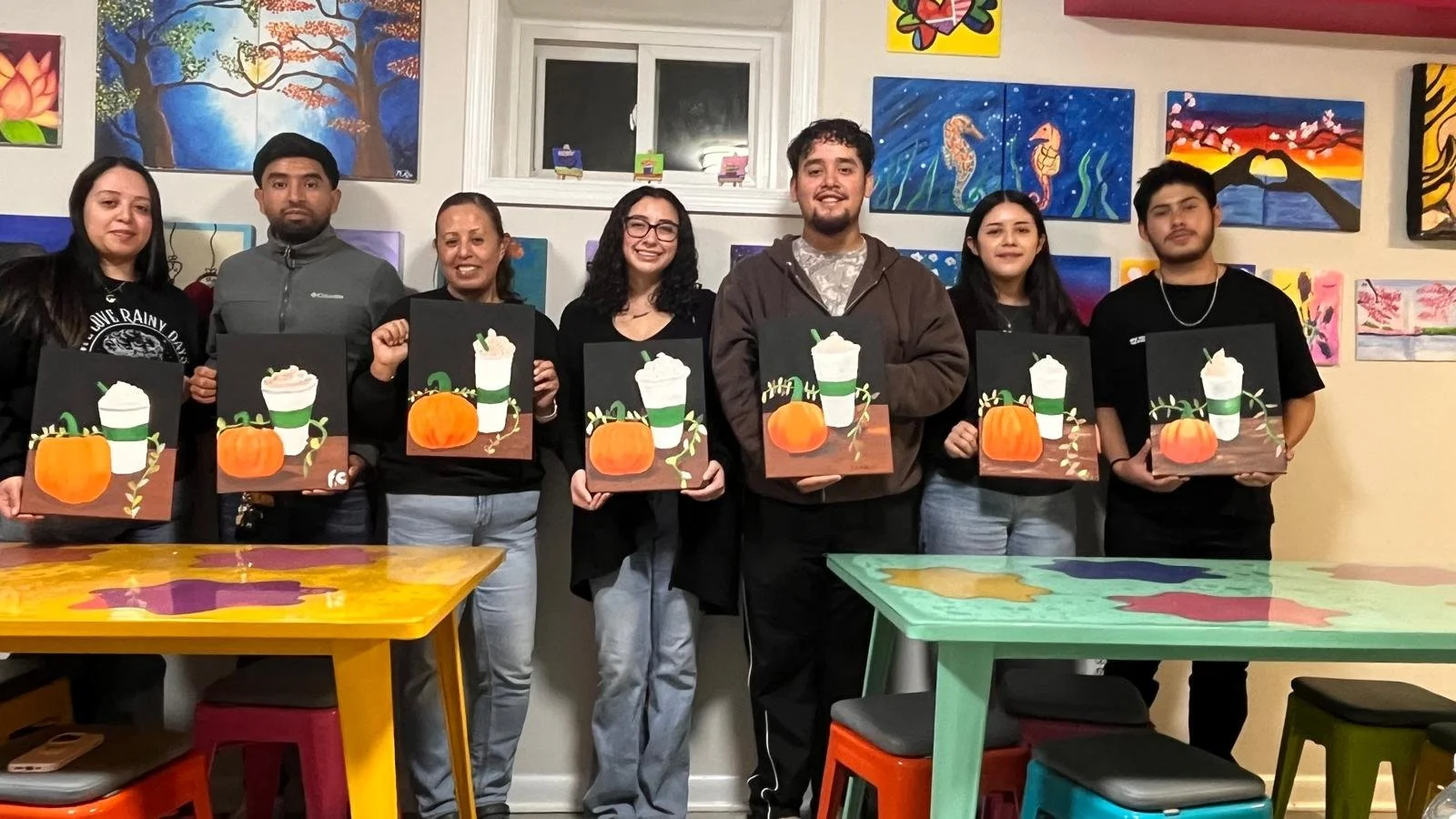 Group of seven people standing in a room holding crafted artwork of a pumpkin and a pumpkin Spice drink, colorful paintings on the wall behind them, and brightly colored tables and stools in the foreground.