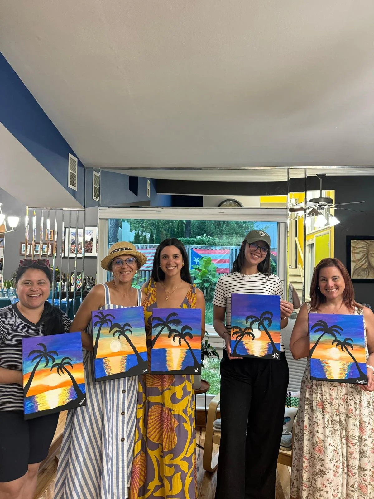 Five women standing in a room holding colorful paintings of a sunset with palm trees and a beach, smiling at the camera.