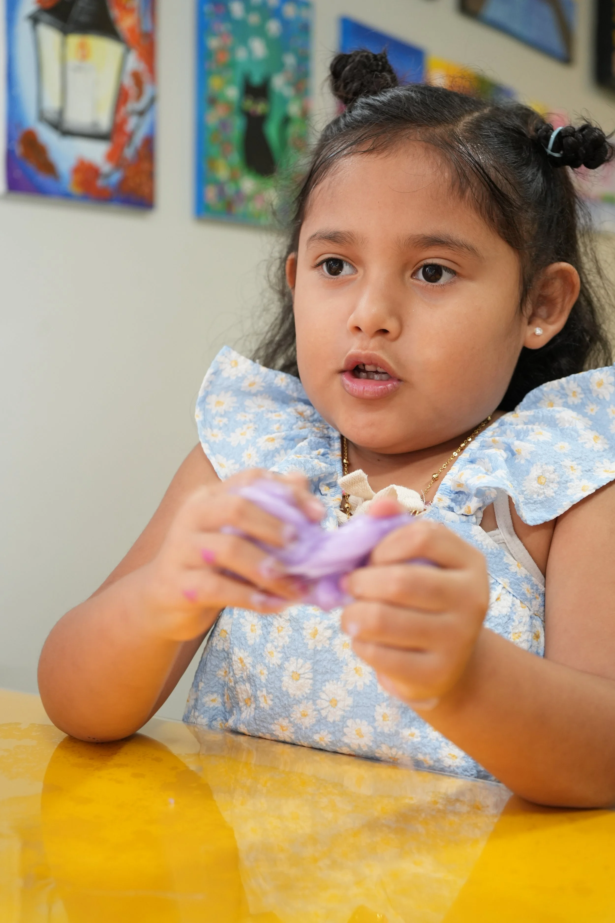 Young girl with dark hair in pigtails, wearing a blue floral dress, sitting at a yellow table, holding a purple bag or object, with colorful artwork on the wall behind her.
