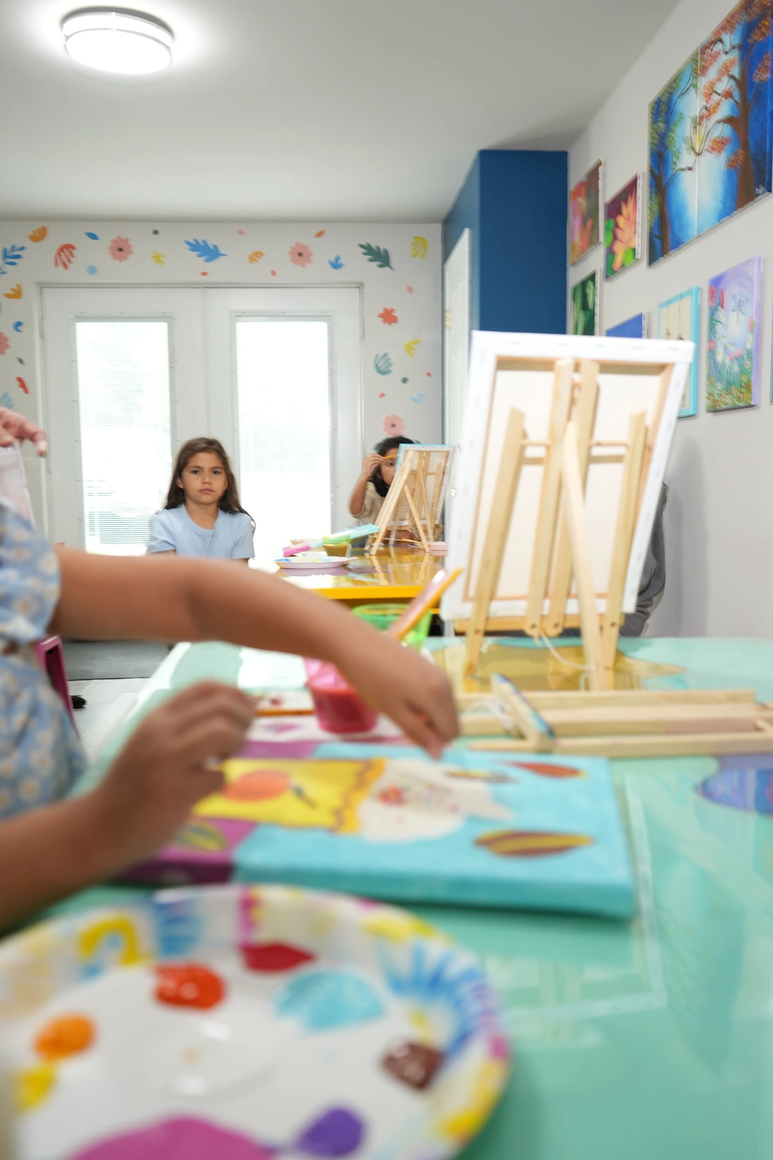 Children participating in a painting activity in an art classroom, with colorful paintings on the walls and easels on the table.