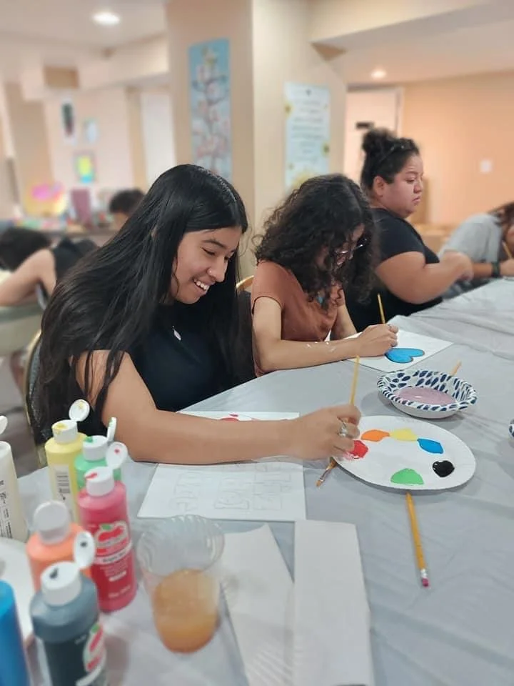 Three young women seated at a table engaged in painting activities, with art supplies including paint bottles, brushes, and colorful paper plates.