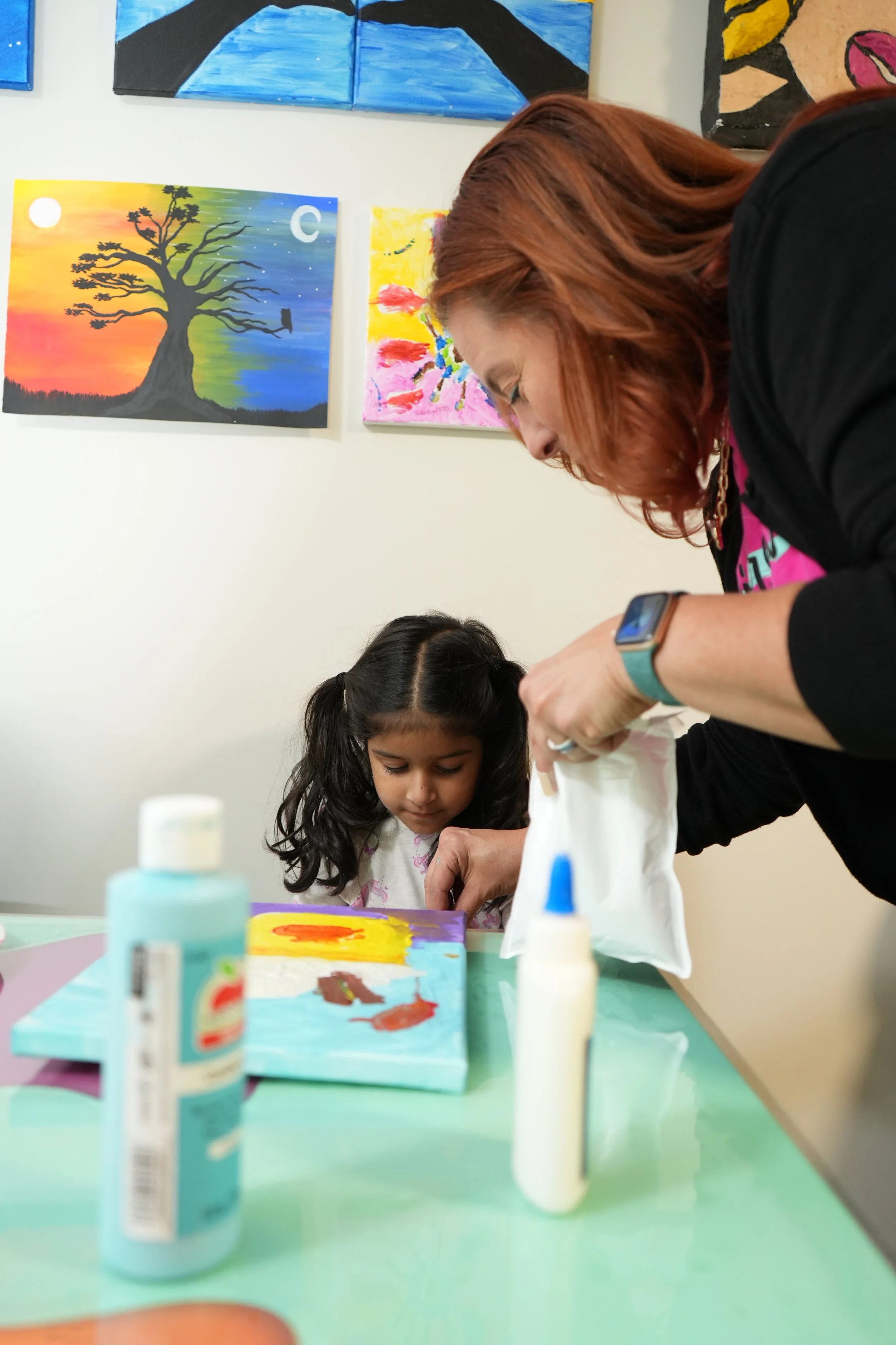 A woman and a young girl engaged in a craft or art project at a table decorated with bottles of glue and painted objects. The woman has red hair and is wearing a black shirt, while the girl has dark hair and is wearing a light-colored shirt. Artworks