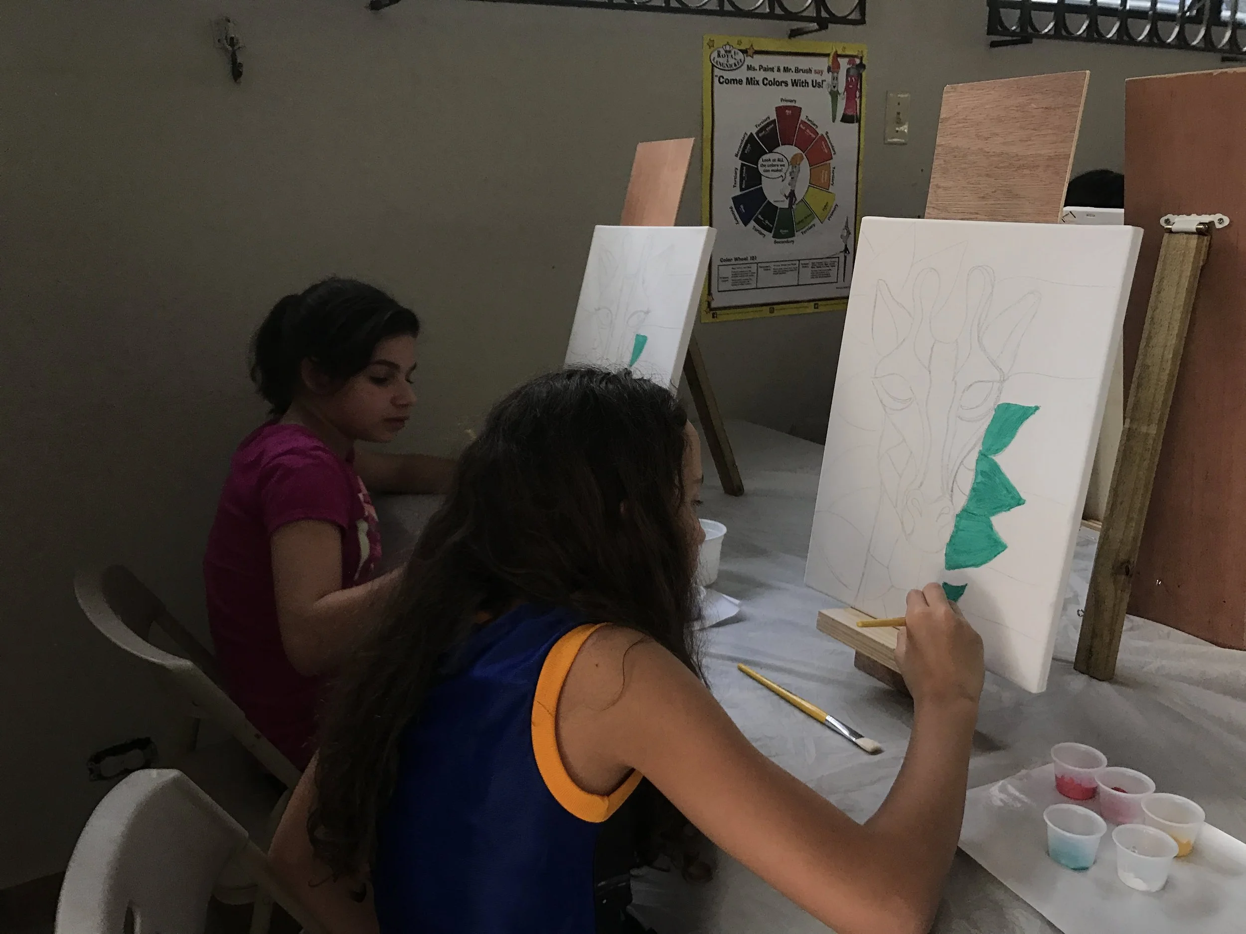 Young girl sitting at a table, painting on a canvas with green paint, surrounded by cups of different colored paints. Other children are also painting nearby, with easels and canvases in the background. A colorful poster about mixing colors is on the