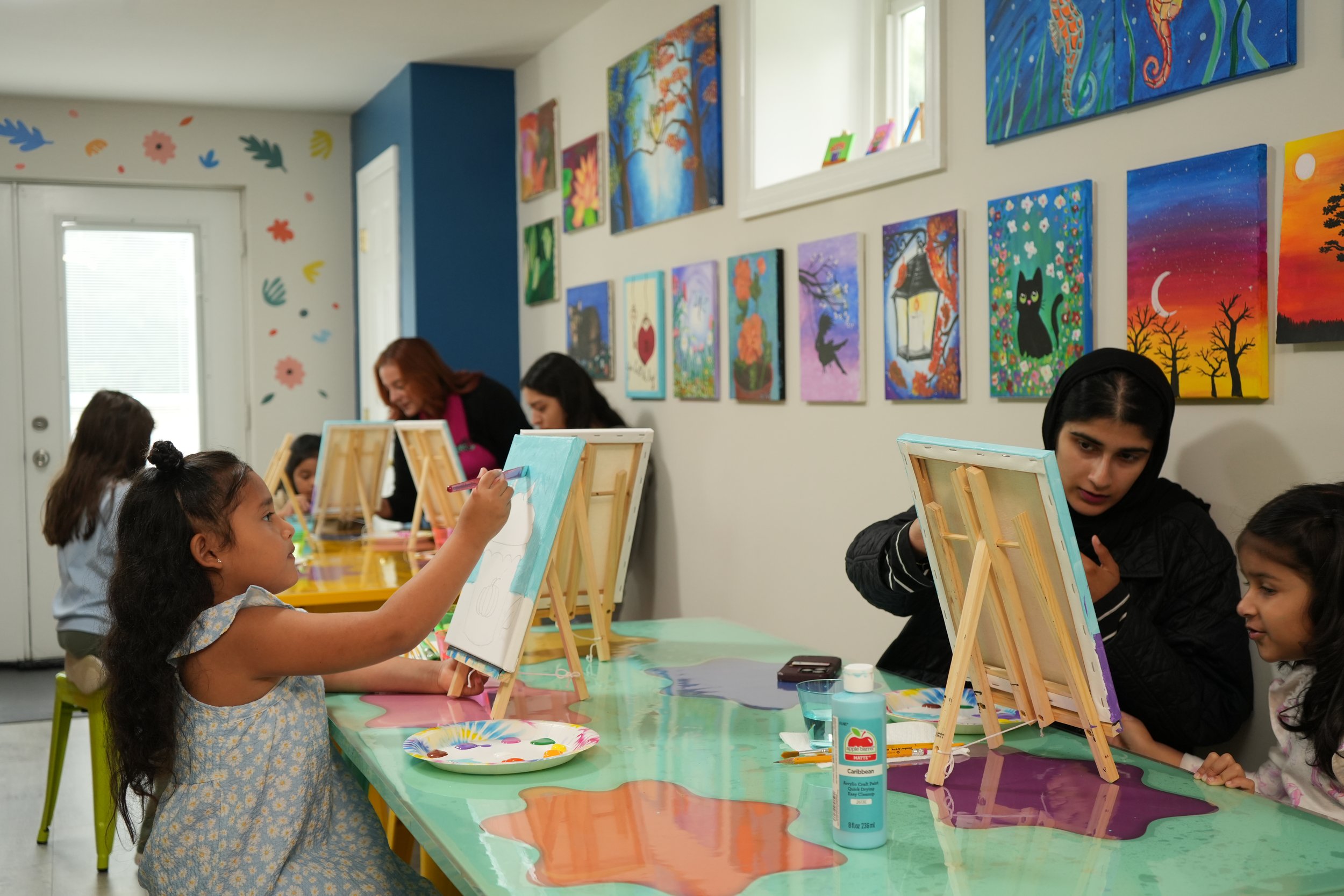 Children and adults participating in a painting class at colorful tables, with framed paintings hanging on the wall behind them featuring various landscapes and whimsical designs.