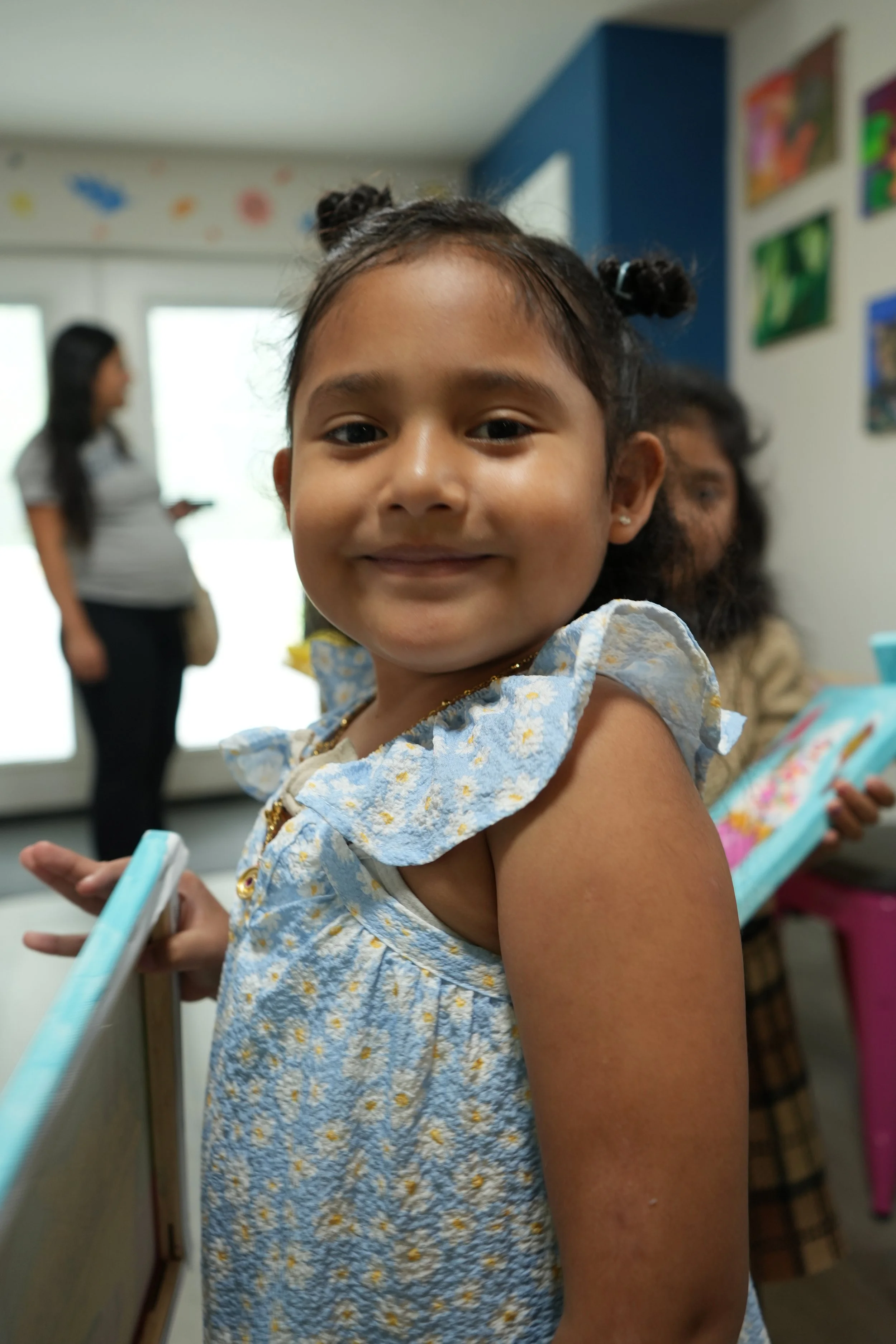 Close-up of a young girl smiling indoors, wearing a light blue dress with yellow and white floral patterns, holding a smartphone, with blurred background of other people and colorful artwork on the wall.