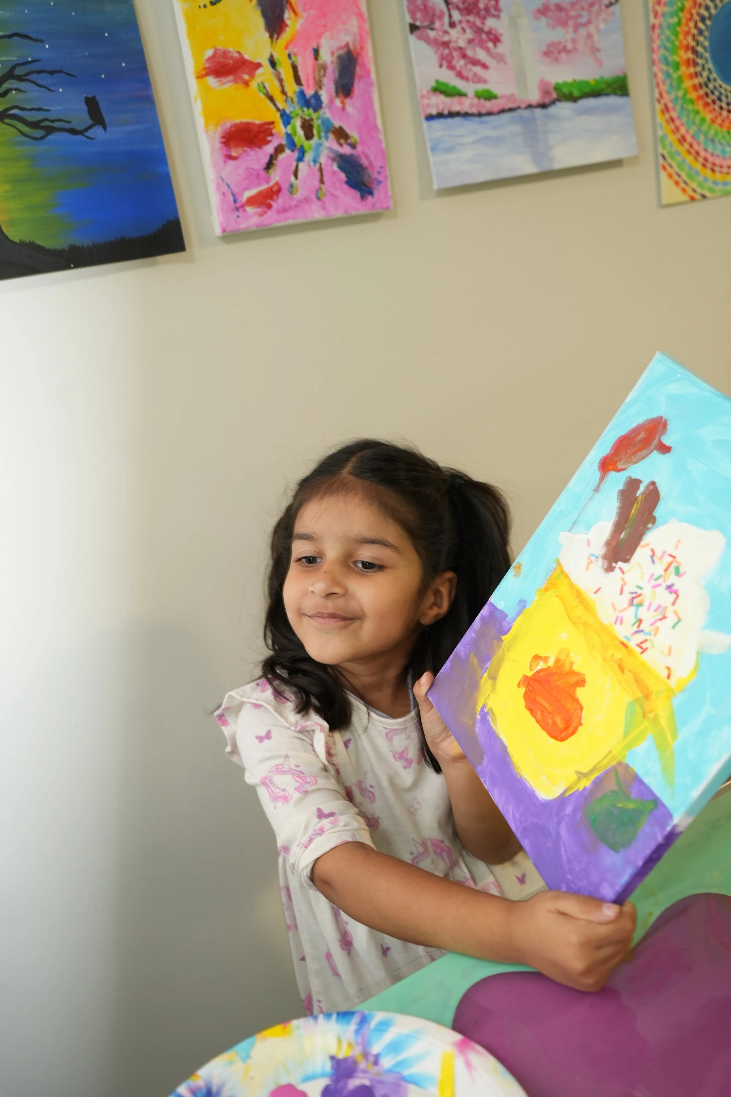 A young girl with dark hair and a white shirt with pink and purple butterflies is smiling slightly and holding a colorful painting of a red fish with rainbow sprinkles on a yellow and purple background. In the background, there are several other colo