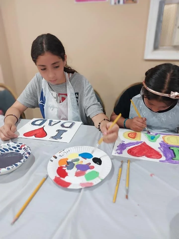 Two young girls sit at a table painting colorful arts and crafts with paintbrushes and paper. They are focused on their art projects, with one girl creating a colorfully decorated sign and the other painting with vibrant colors. The table has paint p