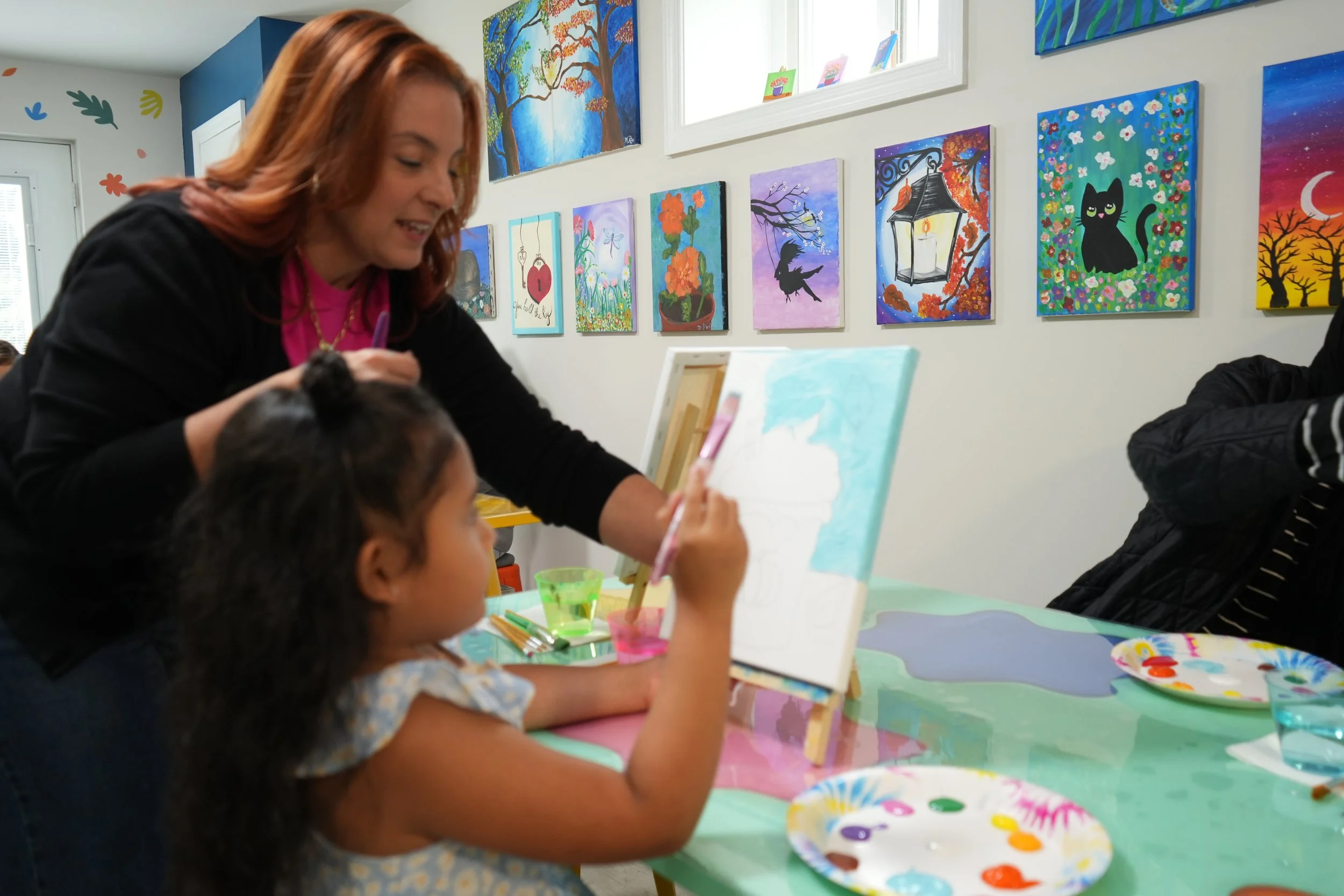 A woman with reddish hair and a young girl with dark hair painting at a table in an art gallery. The woman is smiling and assisting the girl. The scene is decorated with colorful paintings hanging on the wall, featuring trees, animals, and outdoor sc