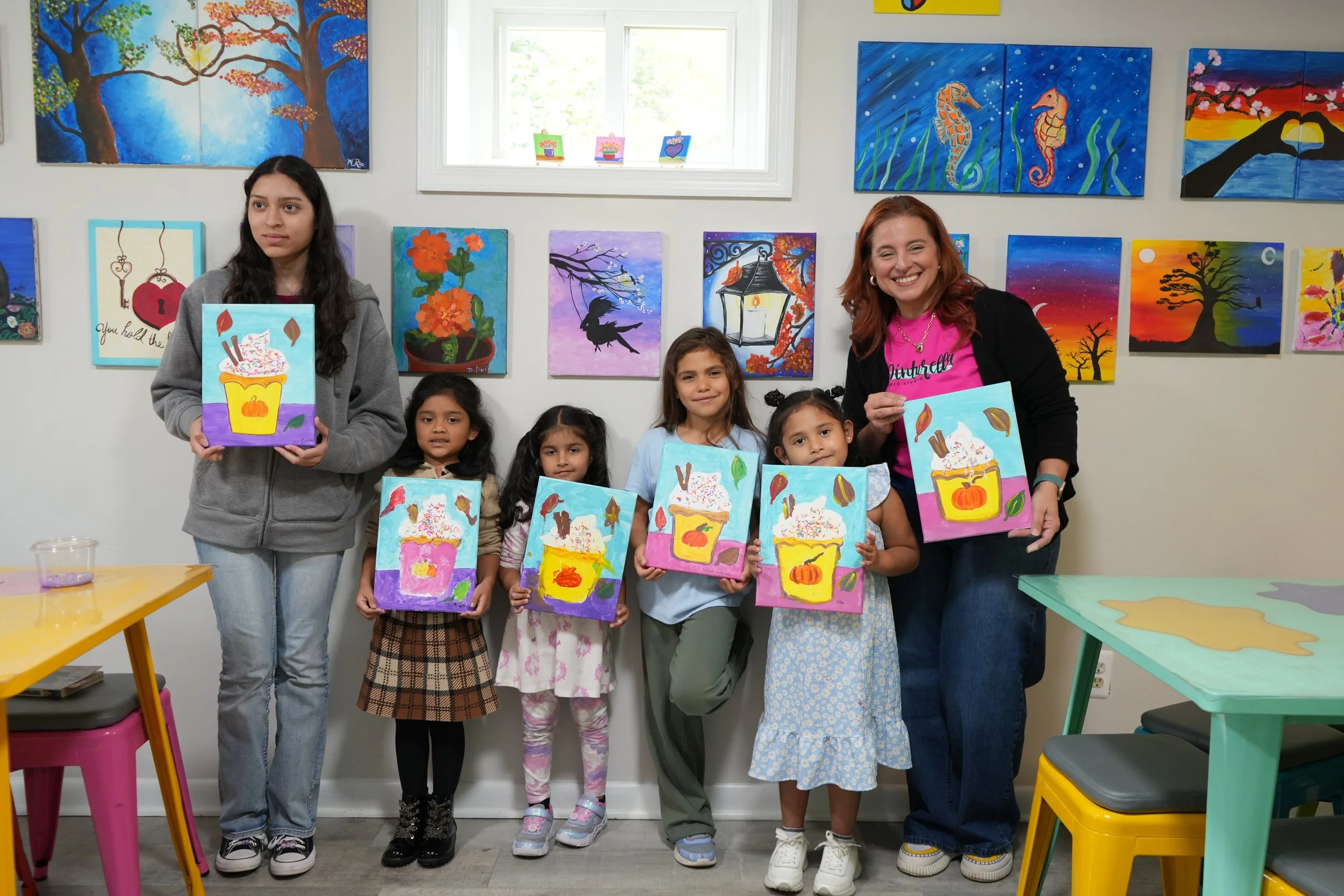 A group of children and two adults in an art gallery holding paintings of a cupcake with colorful frosting, sprinkles, and chocolate sticks. The background features colorful paintings on the wall, including trees, seahorses, birds, and sunset scenes.
