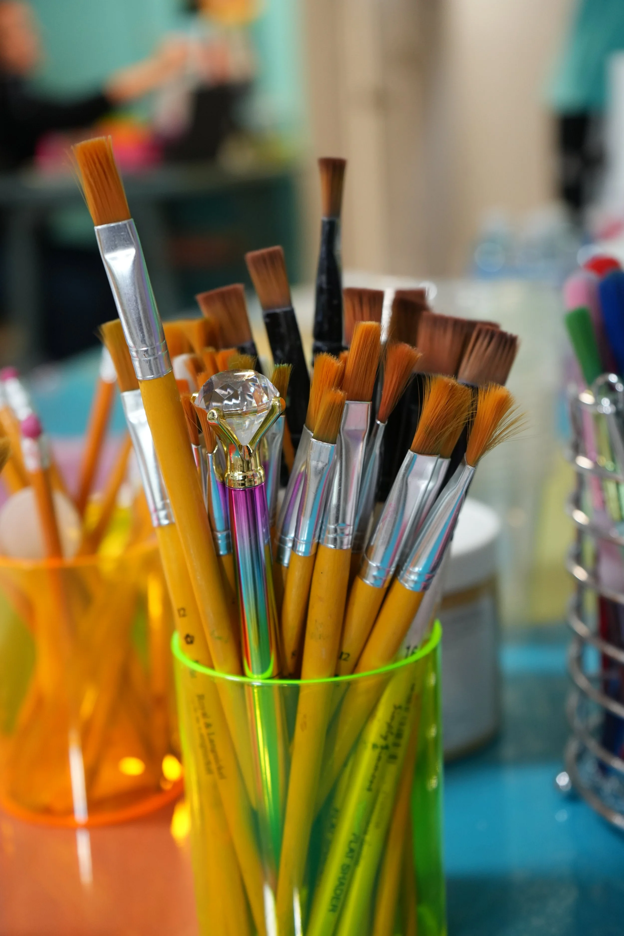 A collection of paintbrushes in various sizes and shapes, stored in a yellow transparent container. Some brushes have wooden handles, and others have black handles, with bristles in shades of brown and orange. A decorative crystal-like knob adorns on