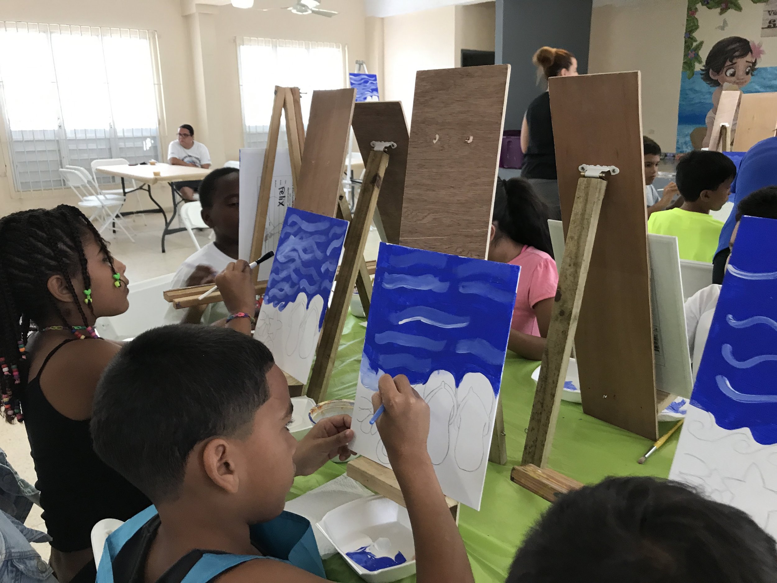 Children painting on canvases set up on easels in a bright room, with a woman assisting. Some children are young and focused on their artwork, which includes blue and white wave designs. A woman with glasses sits in the background at a table.
