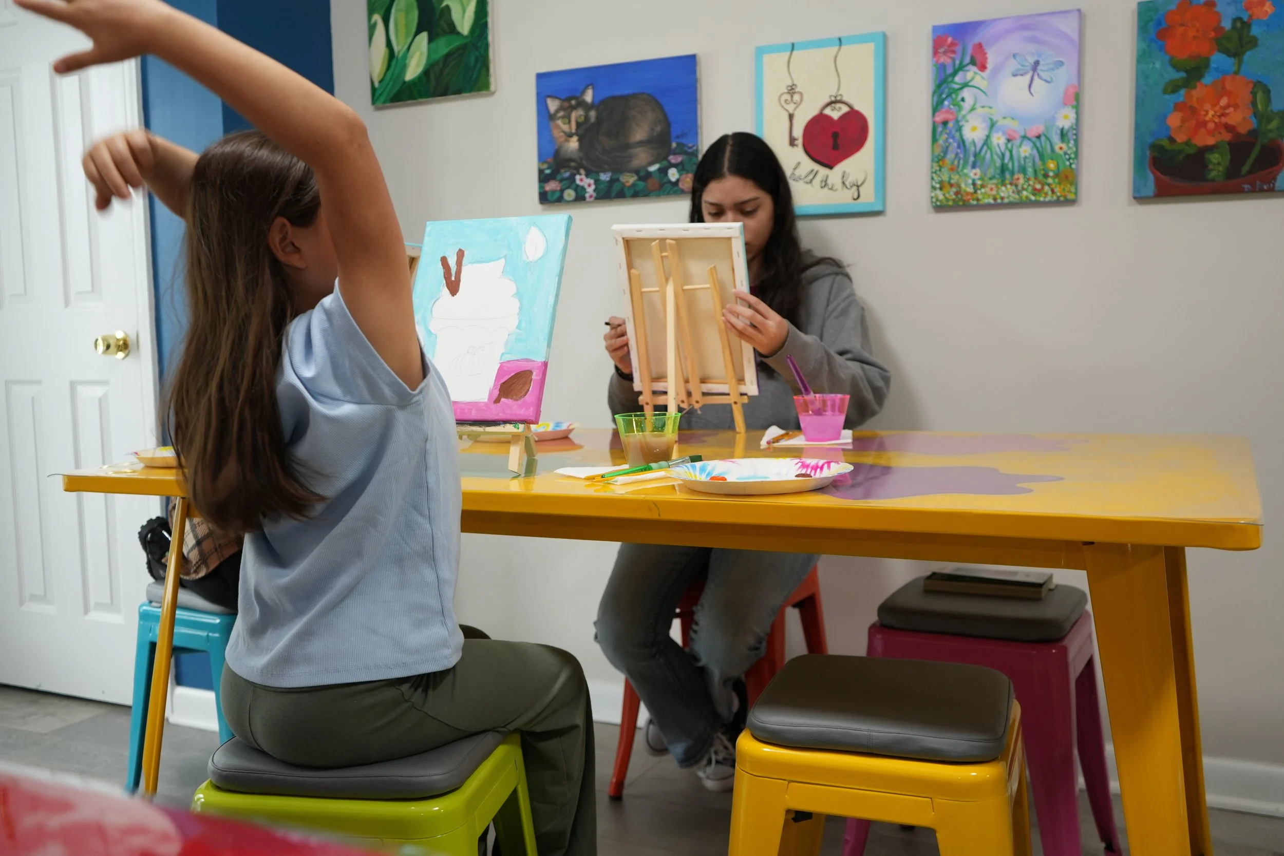 Two girls painting at a table in an art classroom with colorful paintings on the wall.