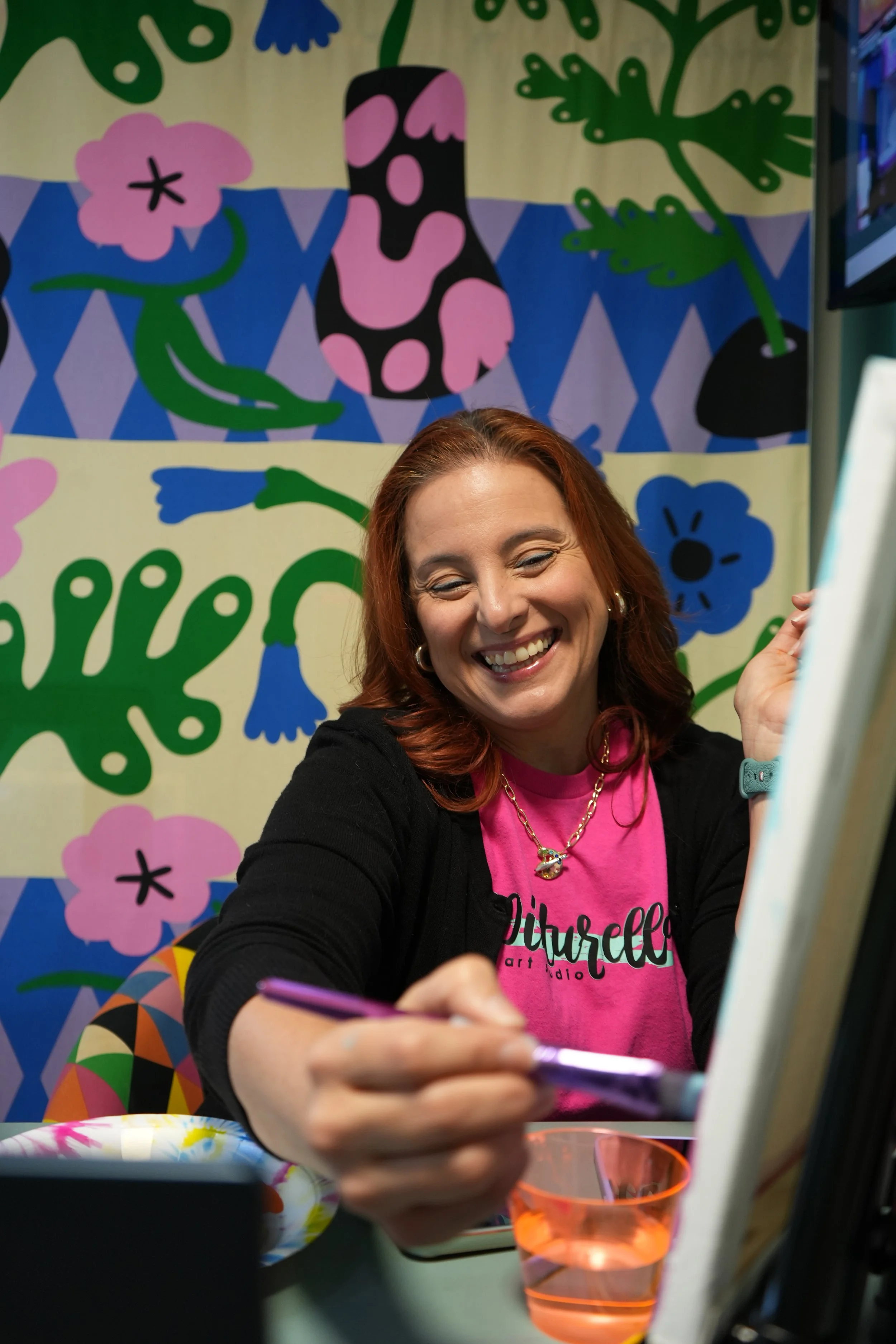 A woman with red hair smiling and writing in a notebook, sitting at a decorated table with a colorful background featuring abstract floral and geometric patterns.