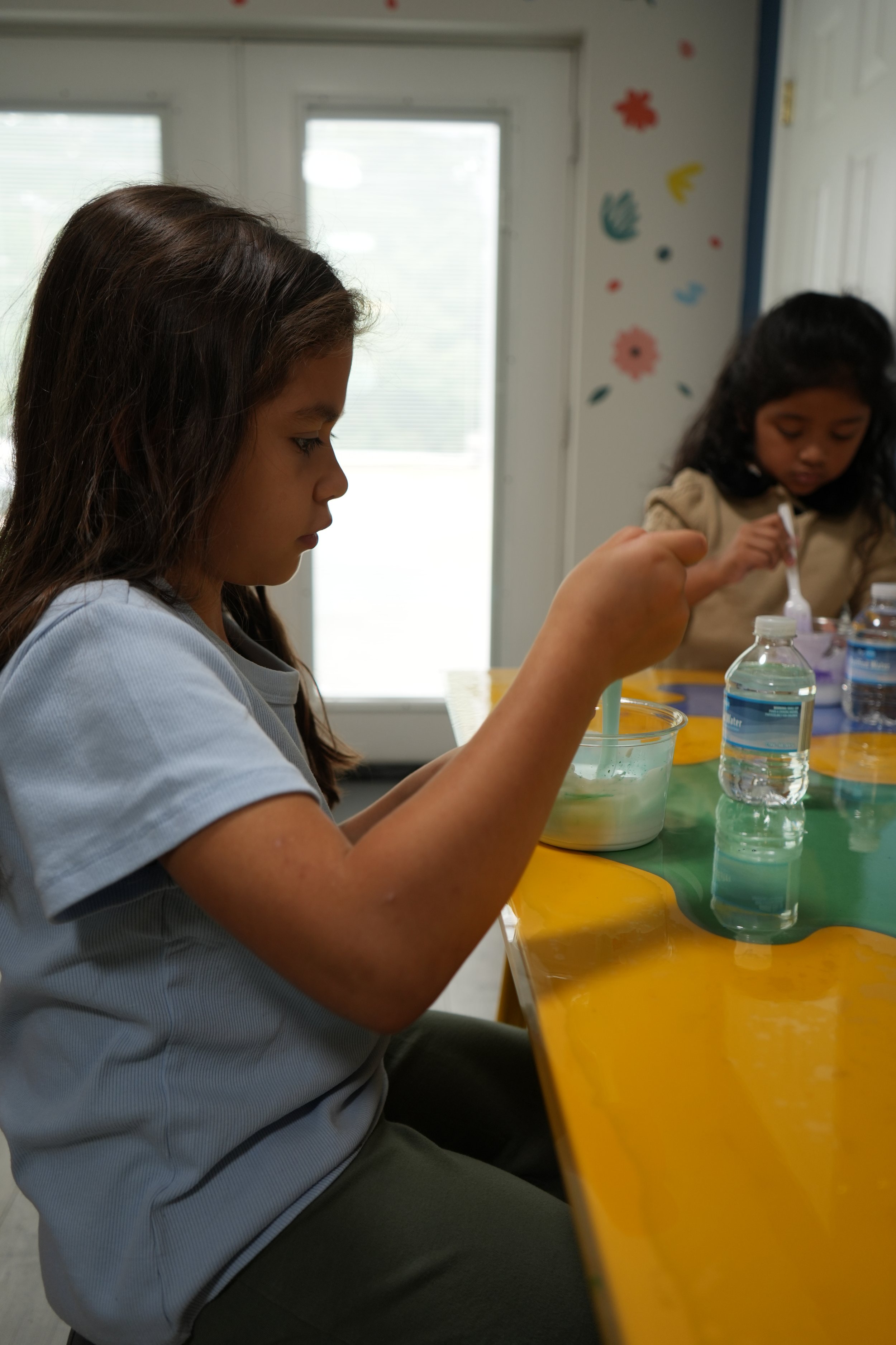 Young girl with long dark hair sitting at a yellow table, stirring a green liquid in a clear bowl, with a water bottle nearby. In the background, other children are engaged in activities and colorful flower and swirl decorations are on the wall.