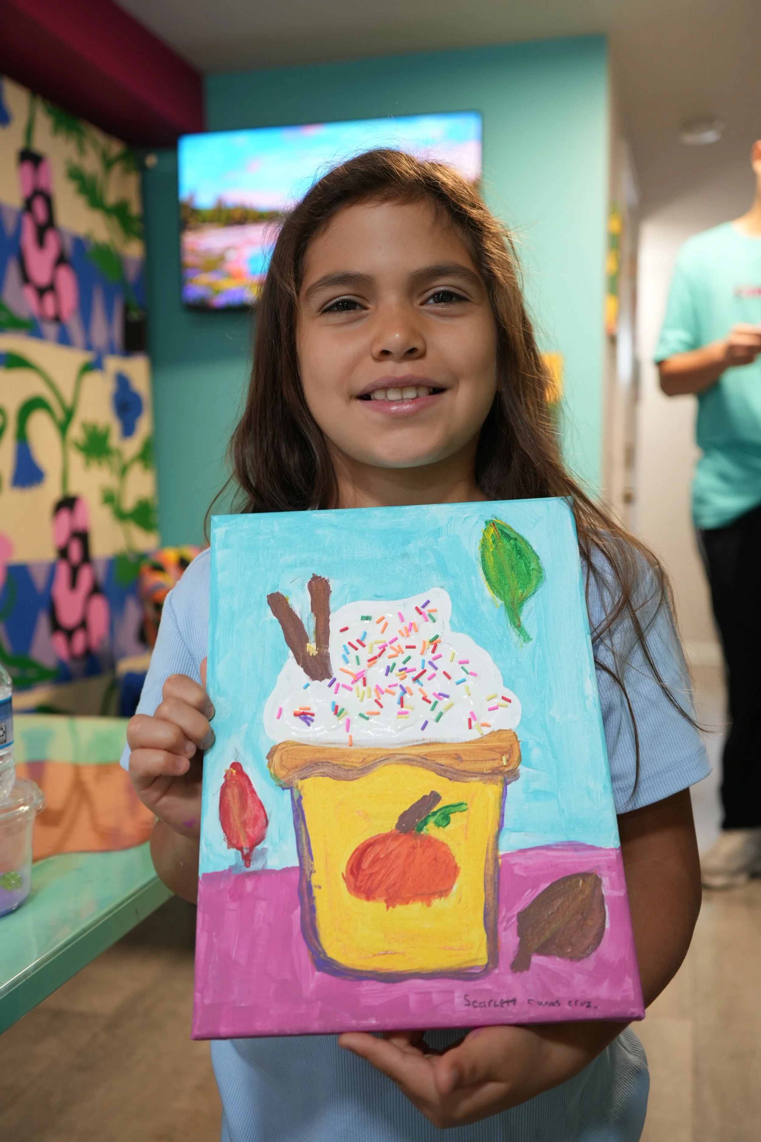 A young girl smiling and holding up a colorful painting of a dessert that resembles ice cream with sprinkles in a glass, a strawberry, and an orange, with a pink background.