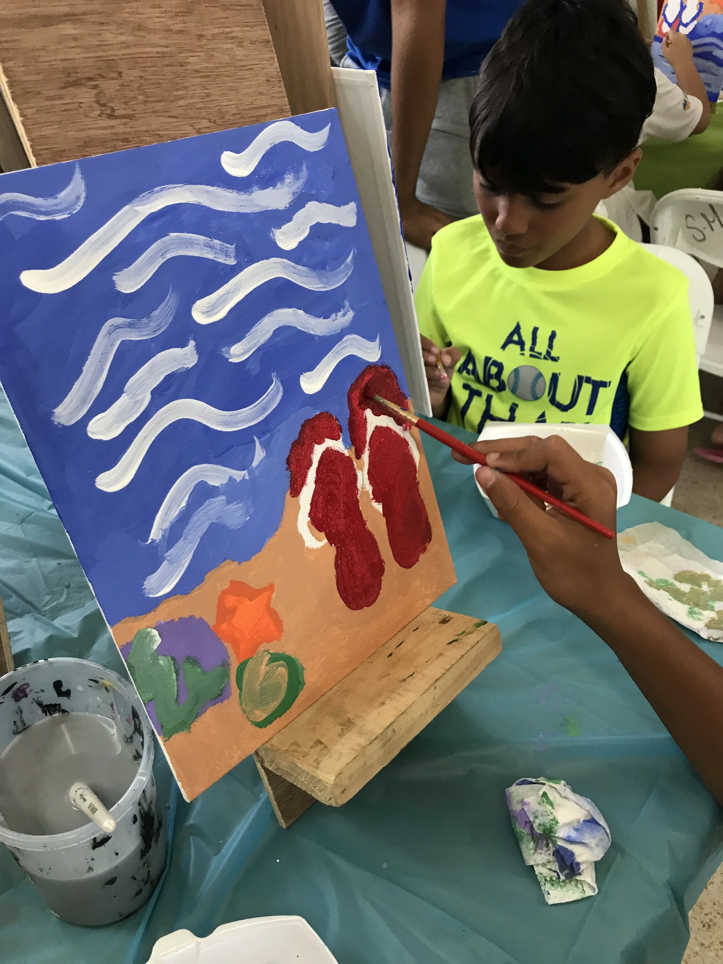 A young boy painting a colorful picture with red, white, and orange shapes on a canvas, at a table with art supplies