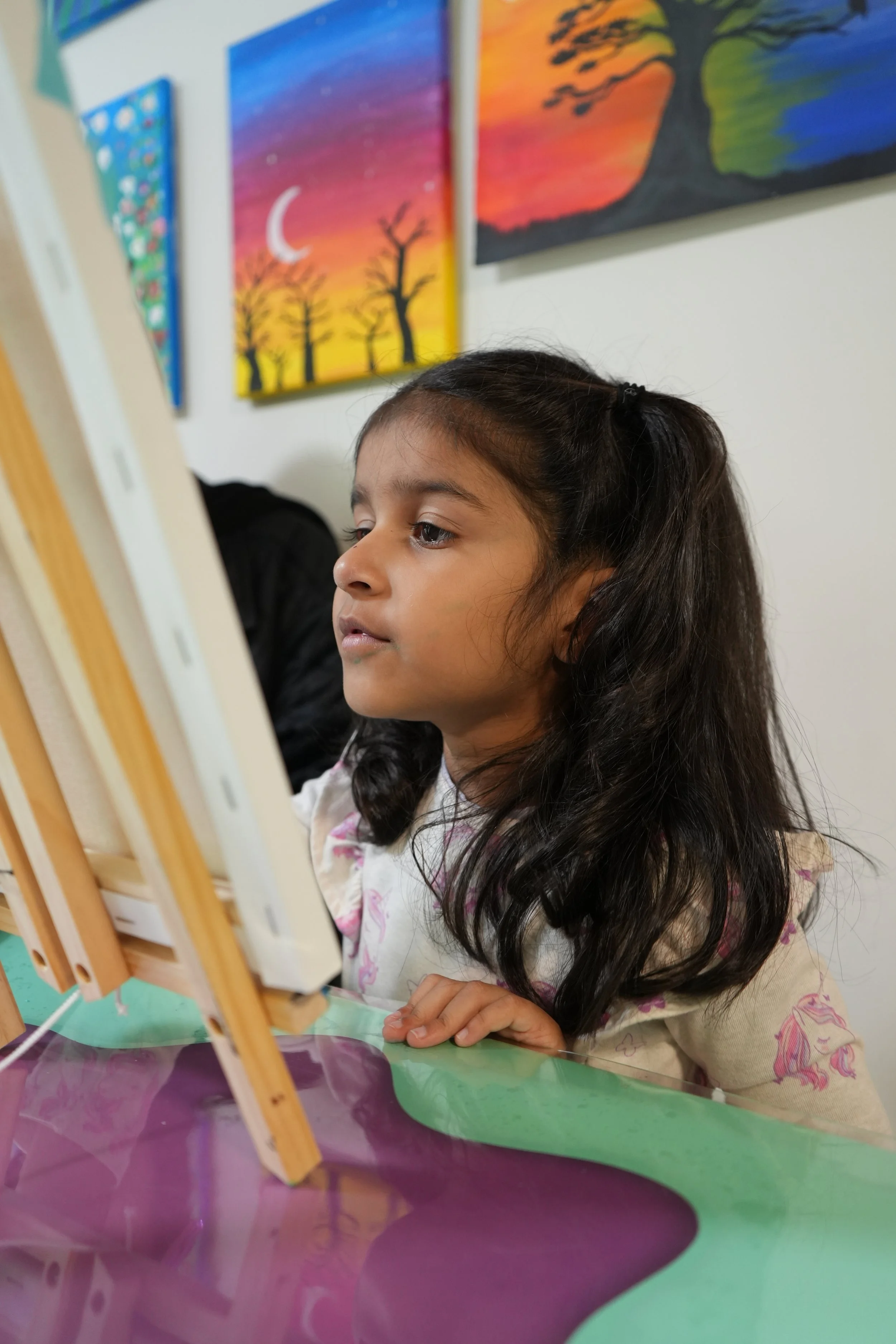 A young girl with long dark hair and a light-colored shirt with a pink and purple design is painting on a canvas inside an art classroom. Her reflection is visible on the purple surface of the table in front of her. Behind her are colorful paintings 