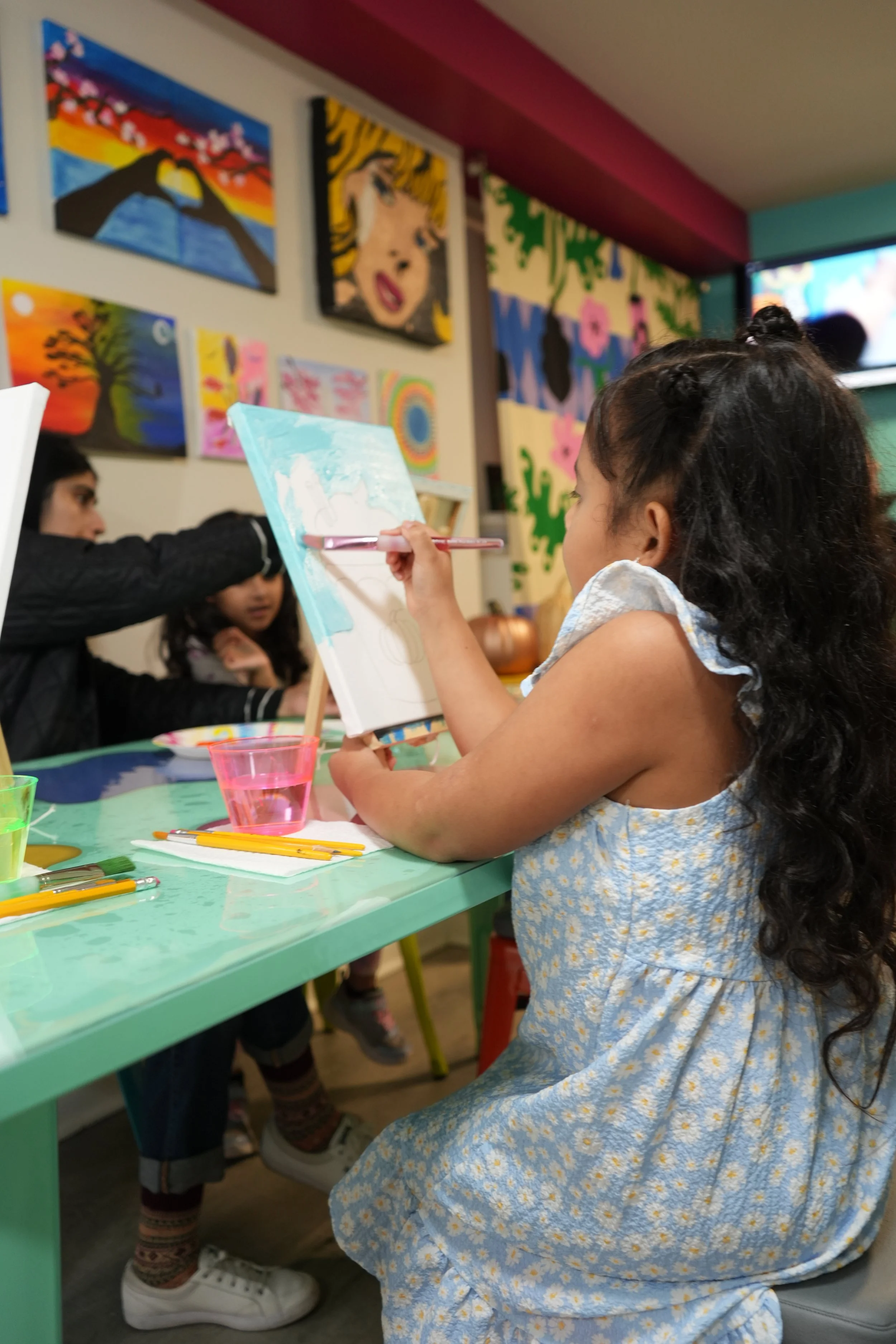 A young girl with long curly hair painting on a canvas at a colorful art studio. She is wearing a blue dress with yellow flowers. Artworks and colorful decorations are on the wall behind her. Other children are visible in the background.