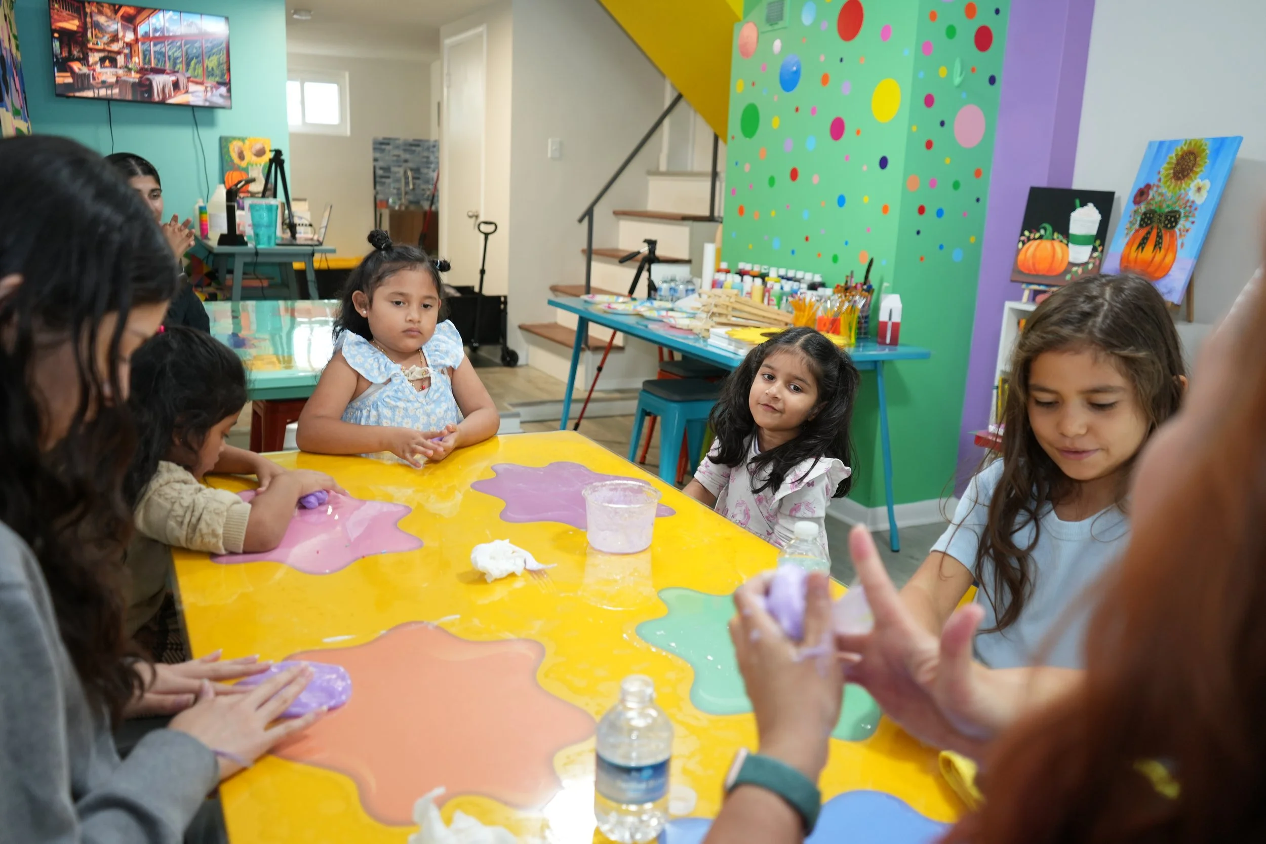 Children participating in a painting activity at a colorful indoor art studio, with art supplies and vibrant wall decorations visible.