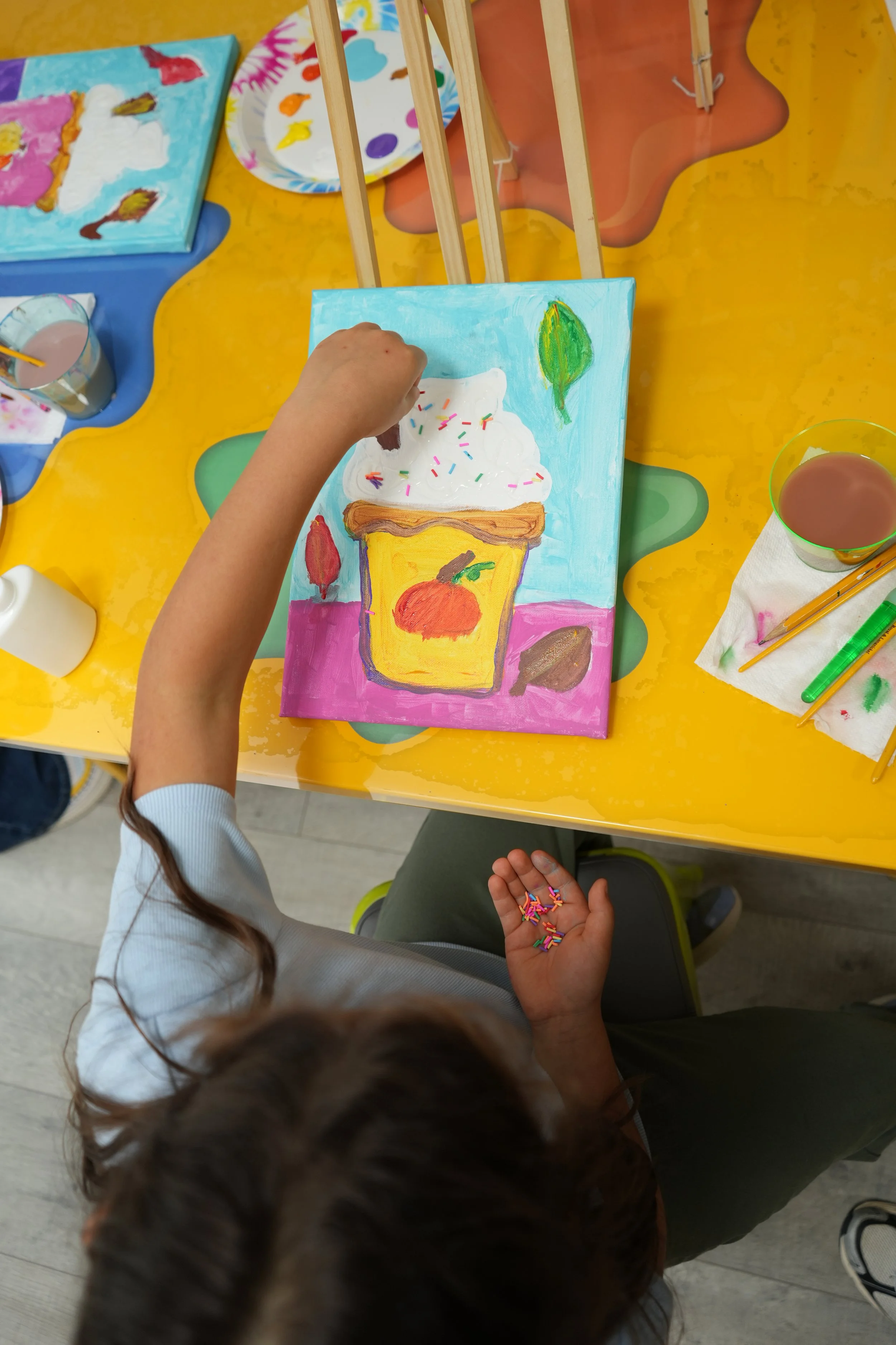 Children painting and decorating a colorful canvas with a cake and fruit design at an art table.