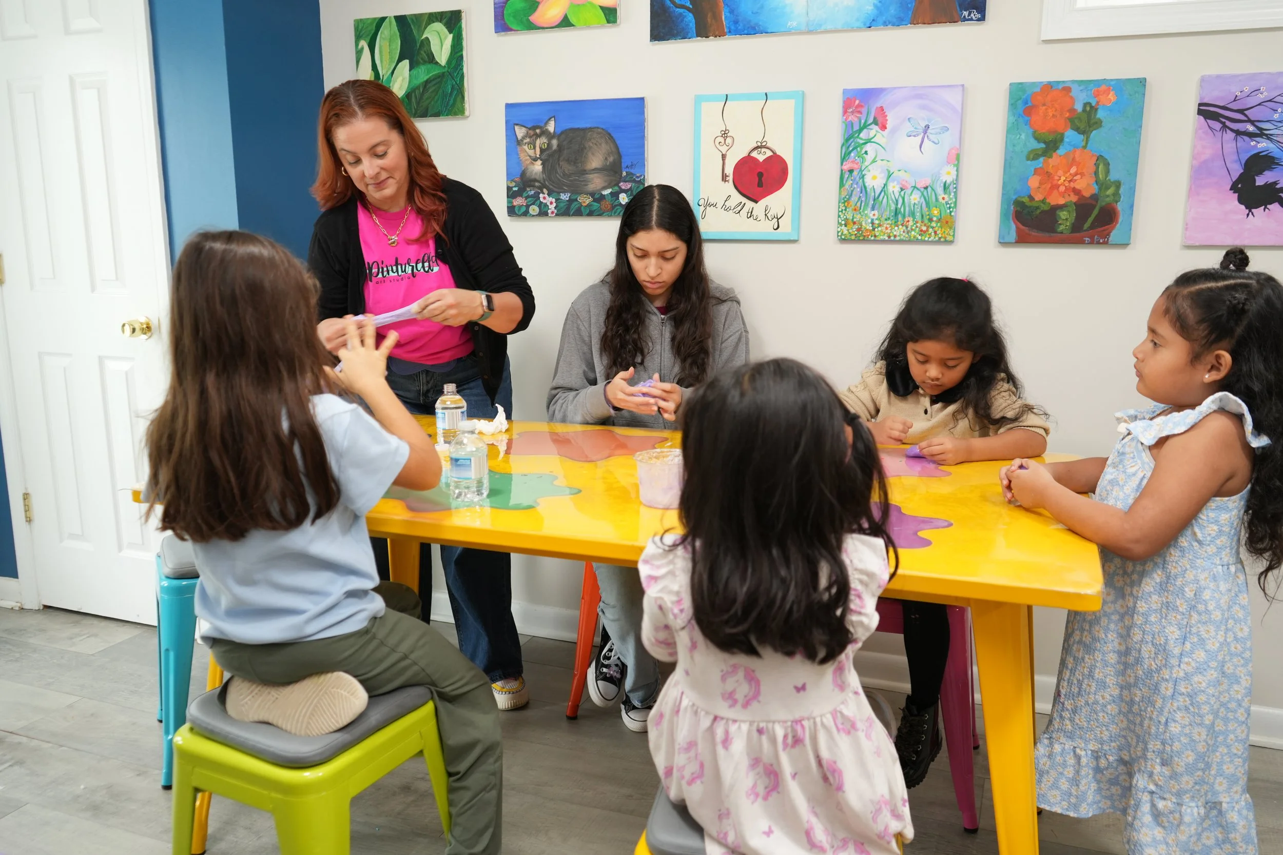 A group of children sitting around a colorful table, engaging in an activity, with an adult woman assisting, in an art-filled room with vibrant paintings on the wall.