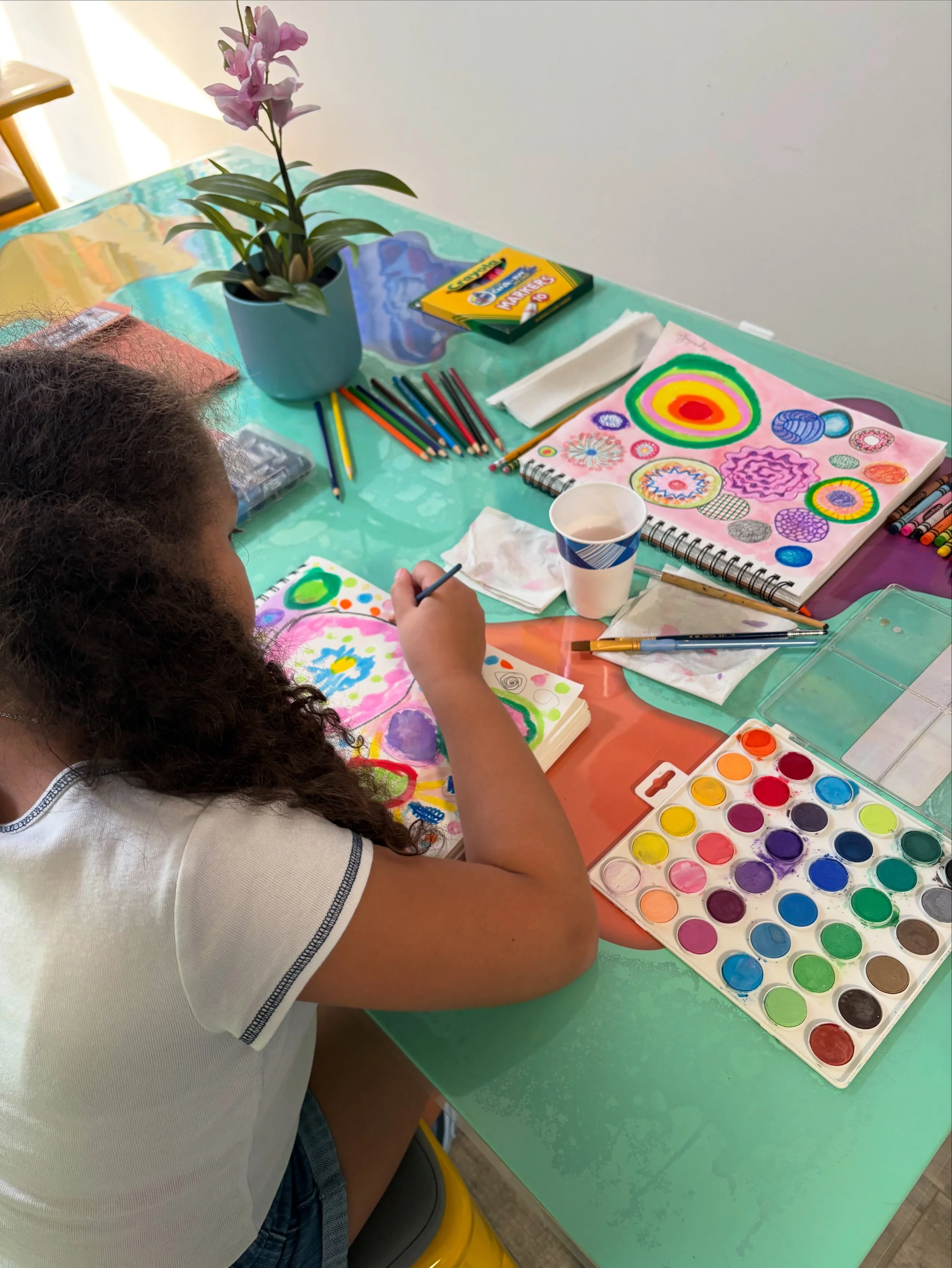 A girl with curly hair painting colorful patterns and designs with watercolor paints on paper, in front of a green table.