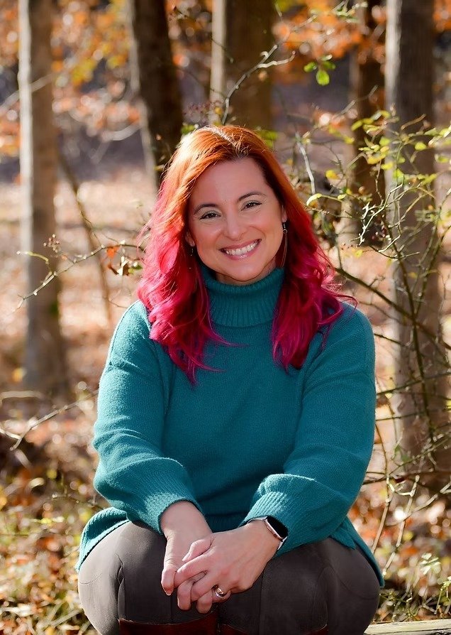 A woman with reddish-pink hair smiling outdoors in a forest, wearing a teal sweater.