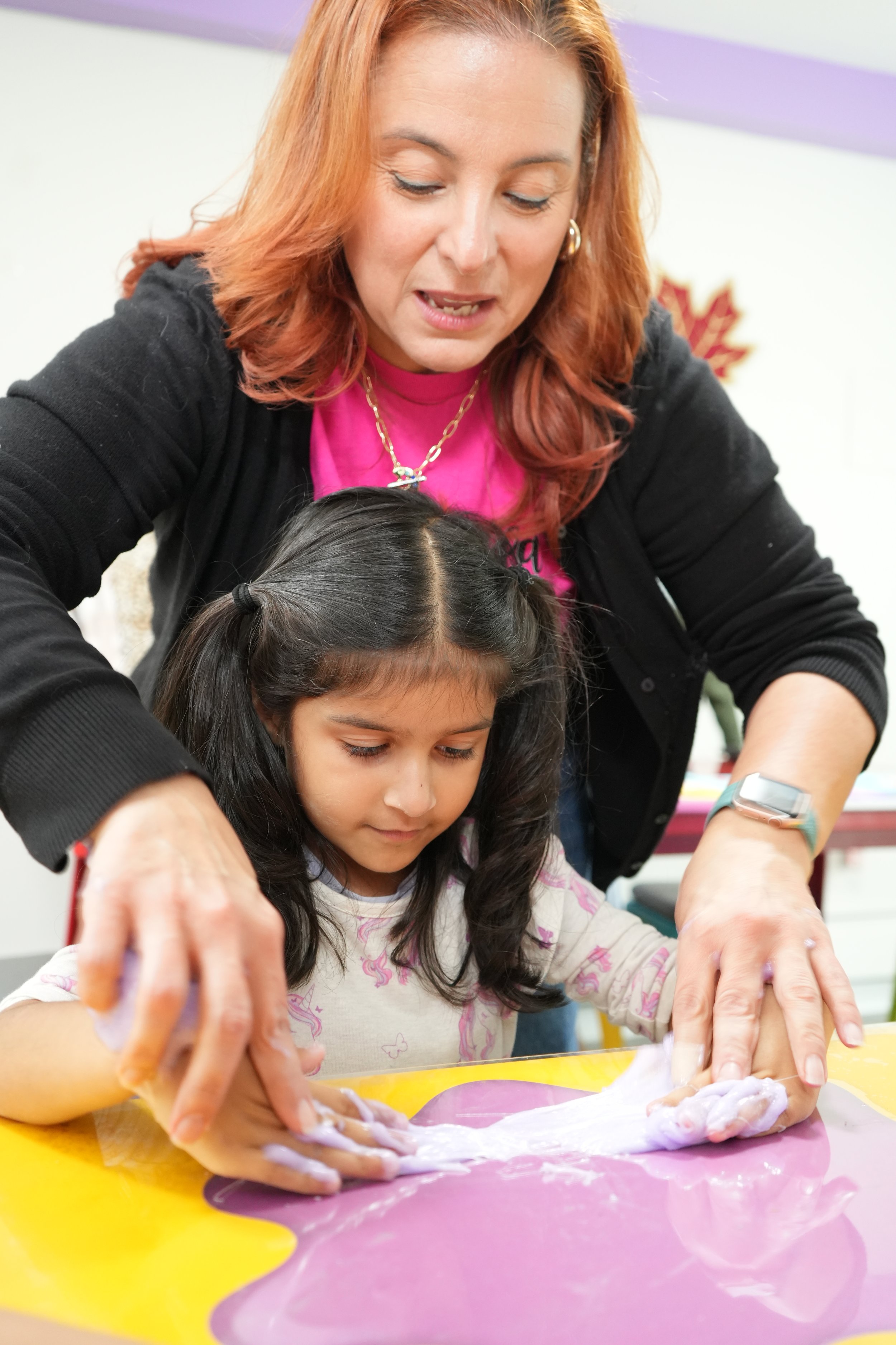 An adult woman with red hair and a child with dark hair are working together on a craft project, kneading a lump of purple slime on a colorful table in a classroom or playroom setting.