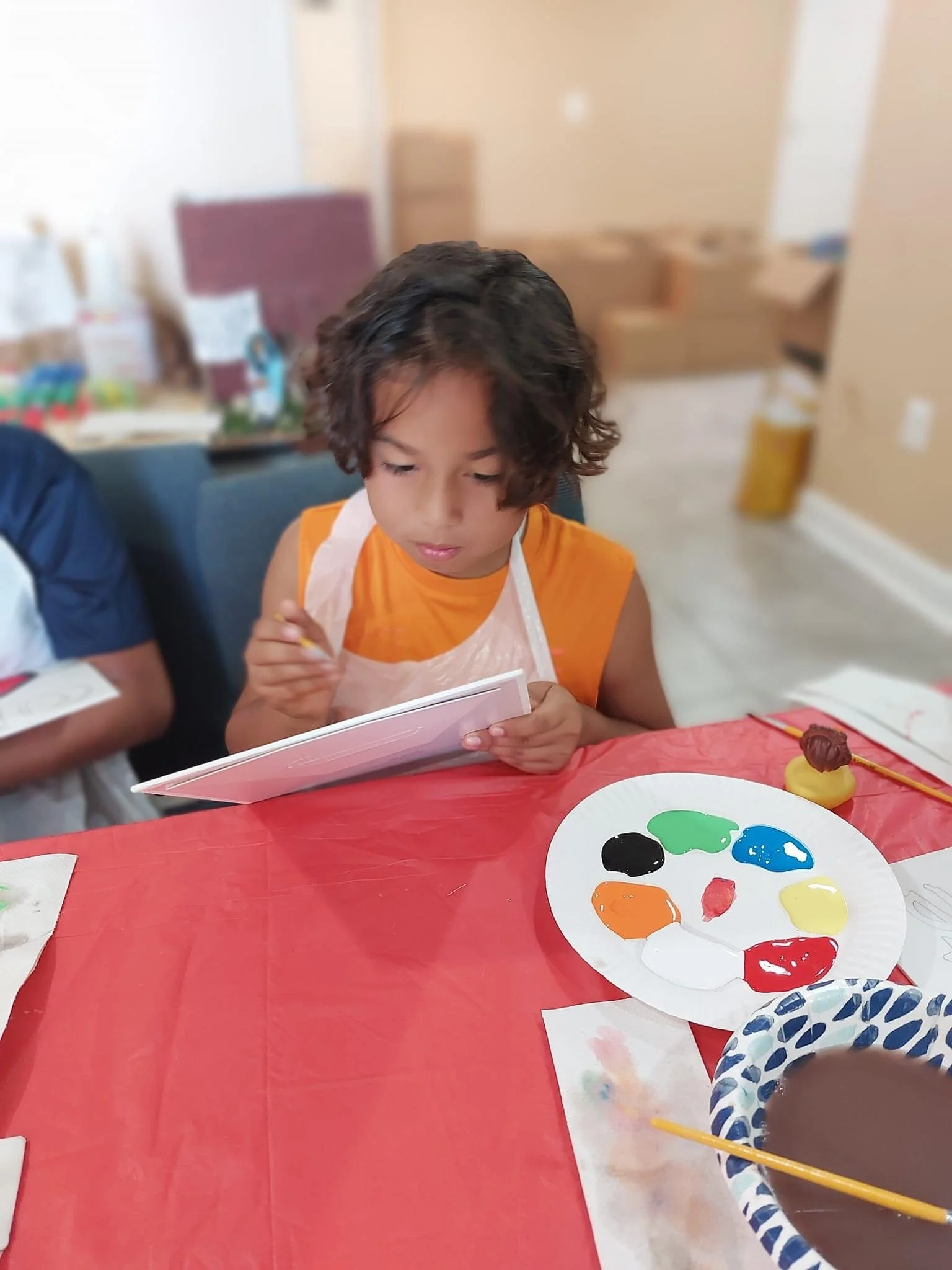 Young boy with curly hair wearing an orange shirt and a plastic apron, painting with various colors on a palette during an art activity at a table covered with a red tablecloth.