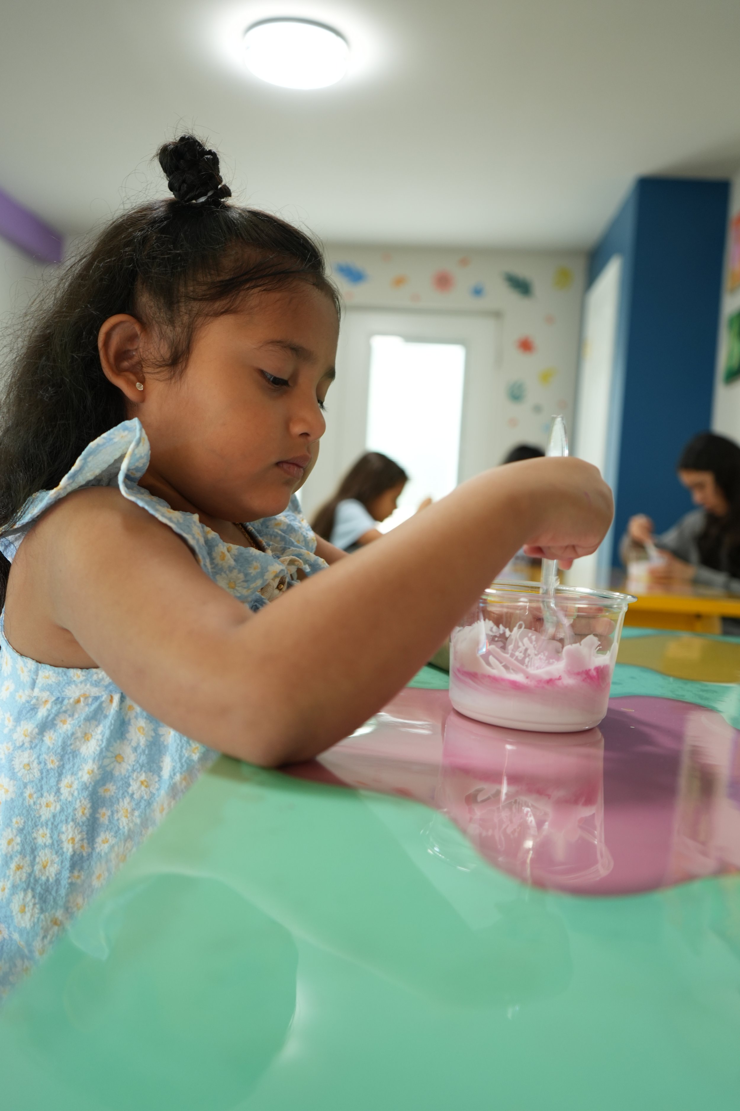 A young girl with dark hair styled in a top knot, wearing a blue dress with yellow flowers, is mixing colors in a clear bowl during an art activity in a colorful classroom.