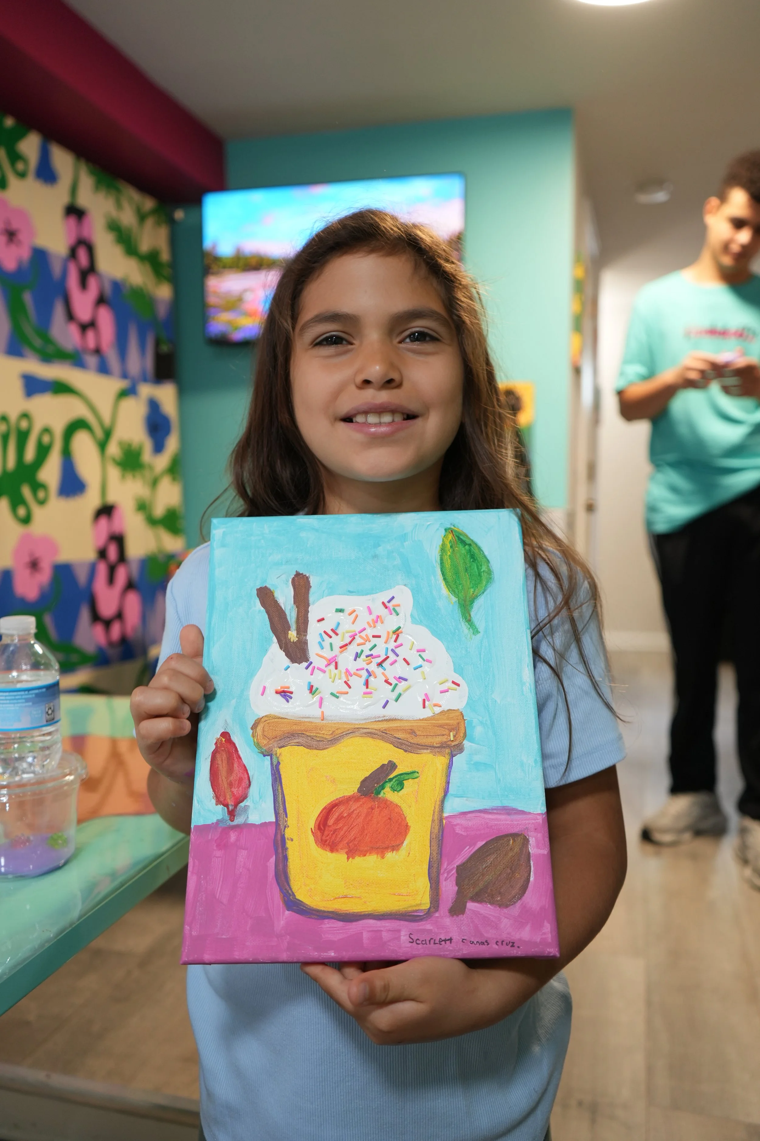 A young girl holding a colorful painting of a dessert with whipped cream, sprinkles, and fruit at a creative art event, with a man in a turquoise shirt and a vibrant mural in the background.