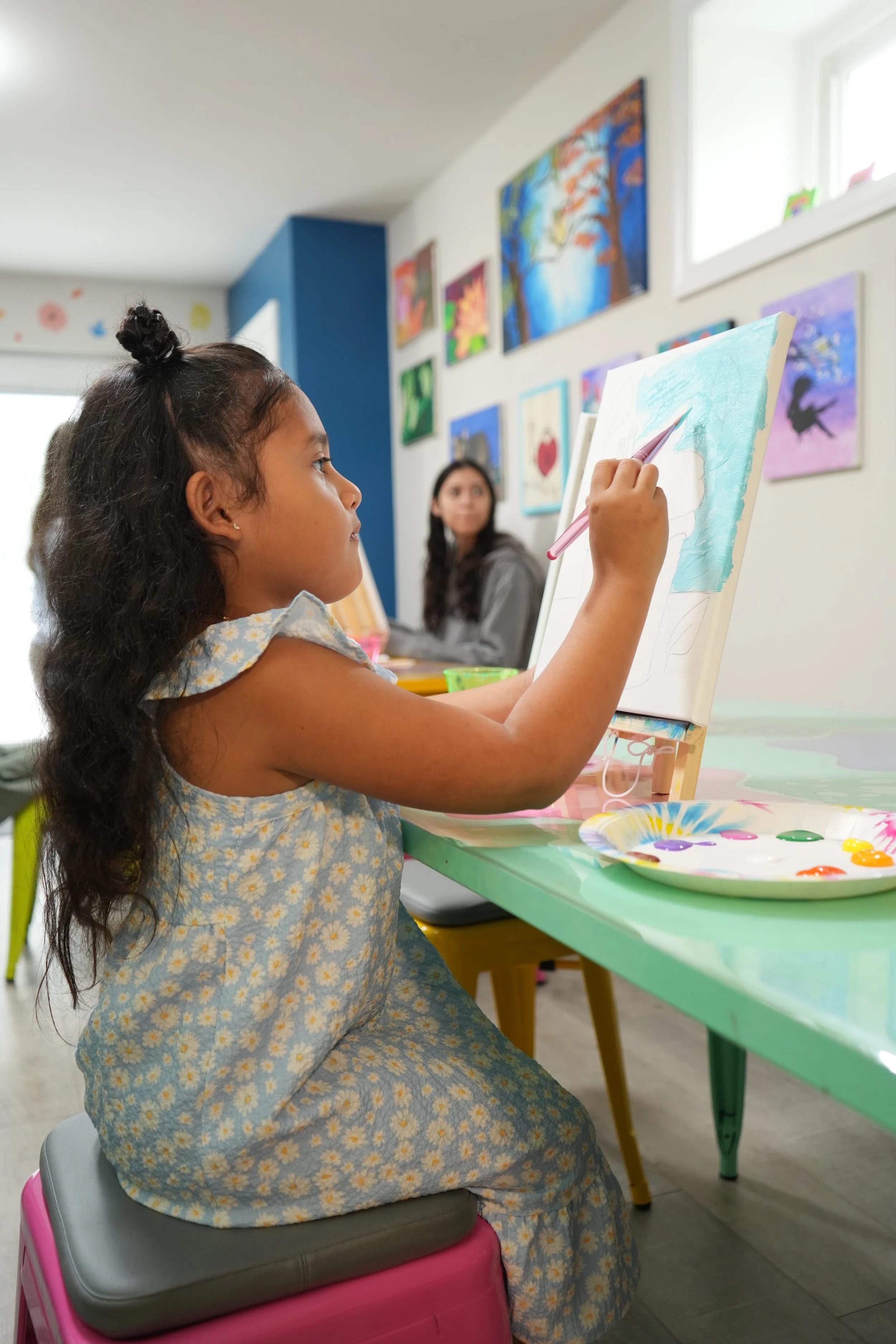A young girl with long dark hair, wearing a blue and white floral dress, is painting on a canvas with blue paint in an art classroom. Another woman, possibly a teacher or parent, is sitting in the background. The classroom walls are decorated with co