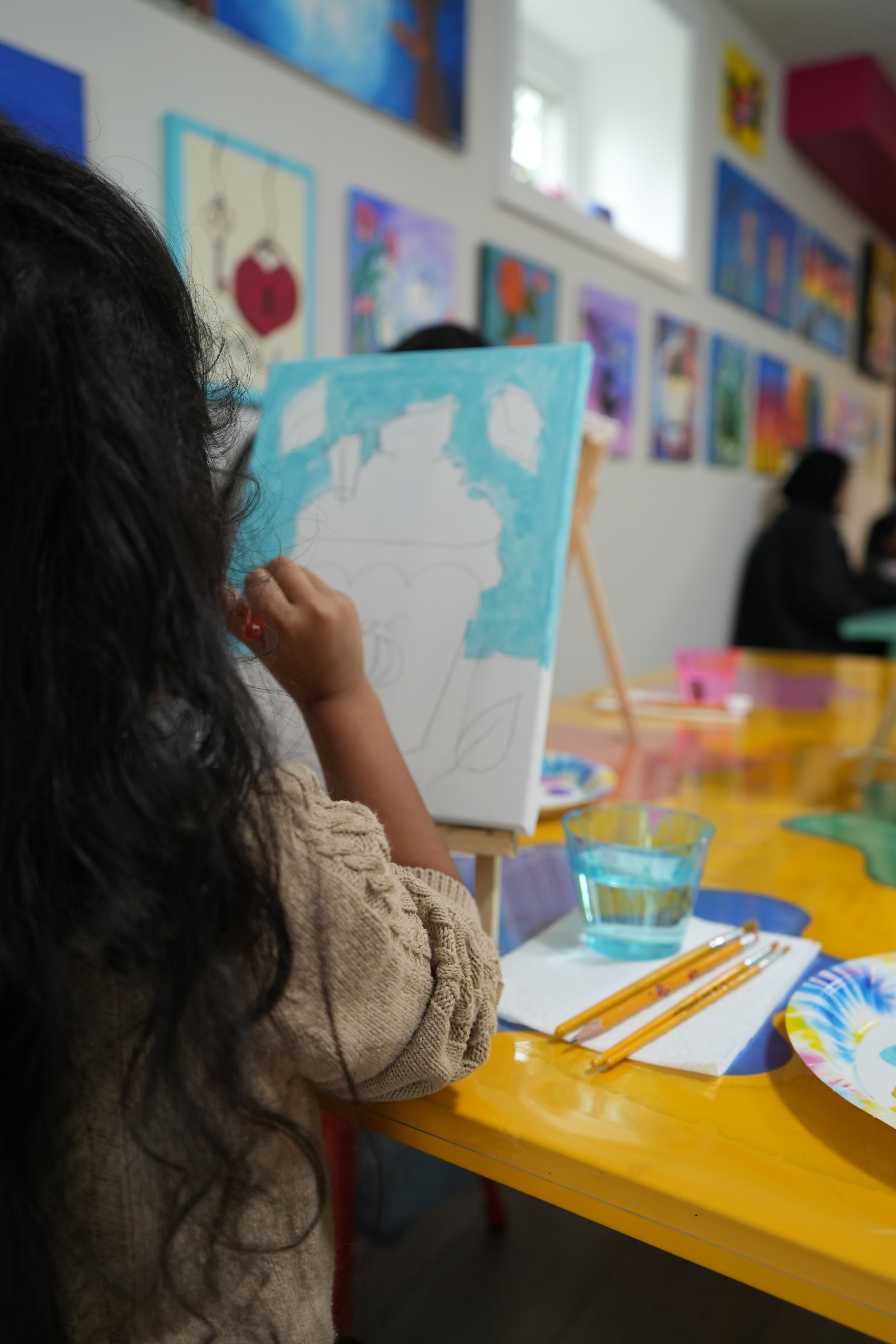 A person with dark, curly hair and a beige sweater is painting on a canvas with a blue background at an art class. The art classroom has colorful paintings on the walls and art supplies on a yellow table, including brushes, a glass of water, and a pa