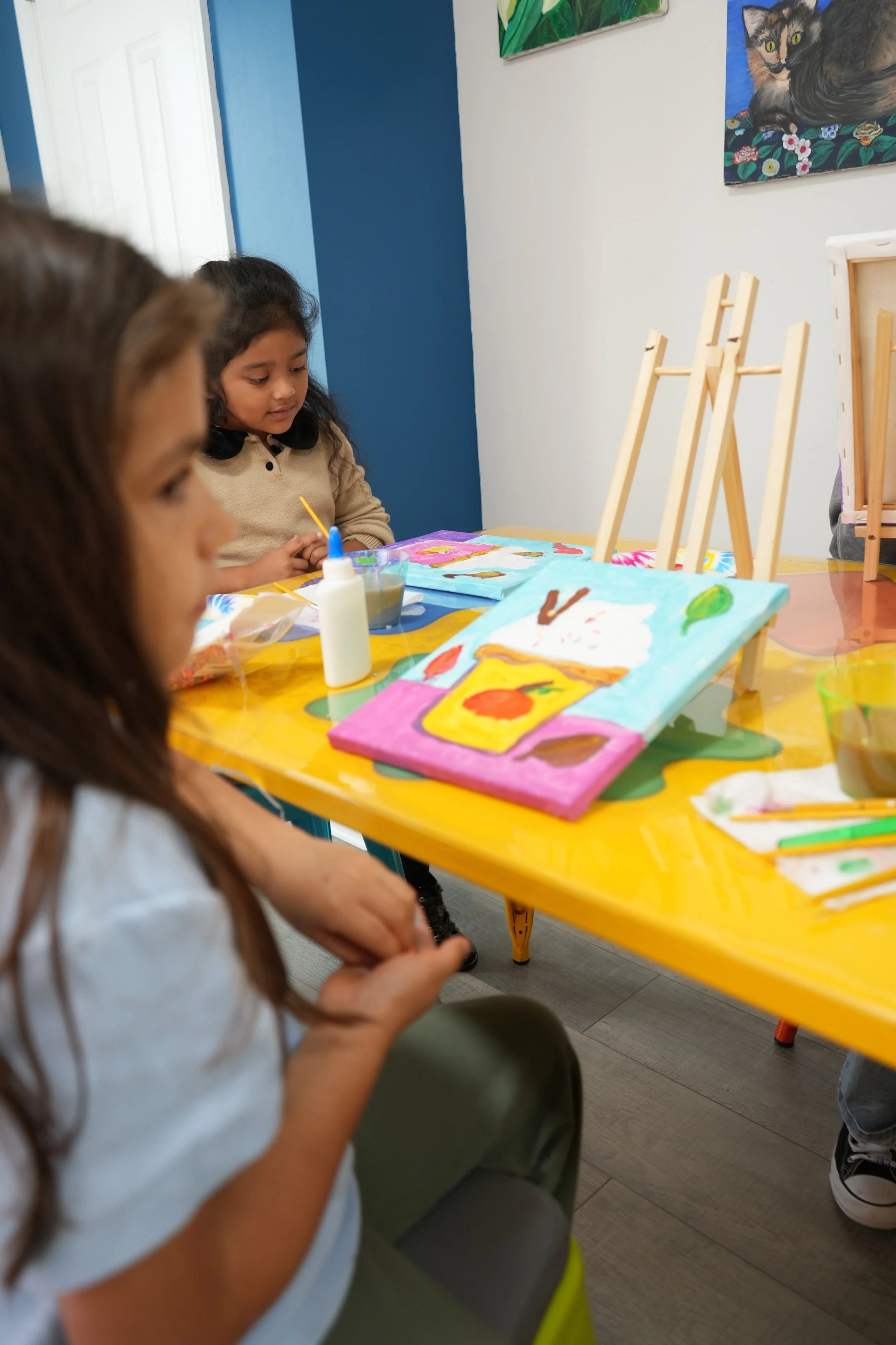 Children painting and decorating with paint and brushes at a bright yellow table in an art room with colorful paintings on the wall.