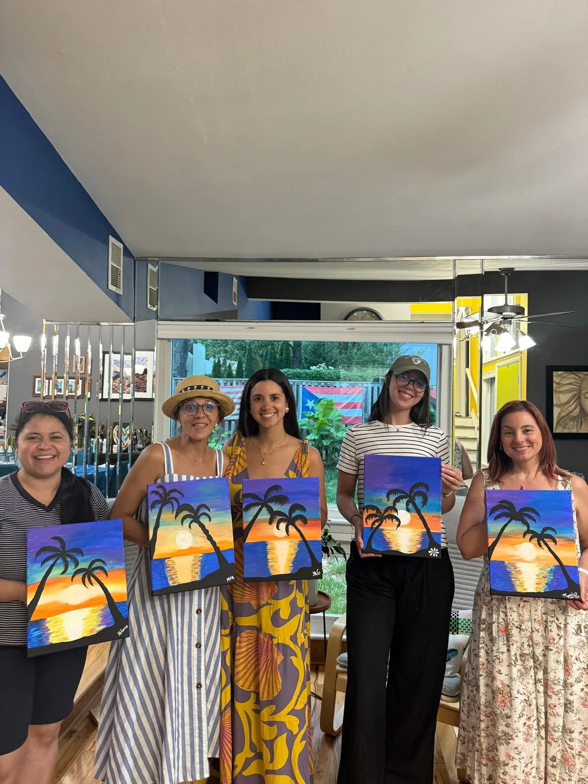 Five women standing indoors, each holding a painting of a tropical sunset with palm trees and water, during a painting event or class.
