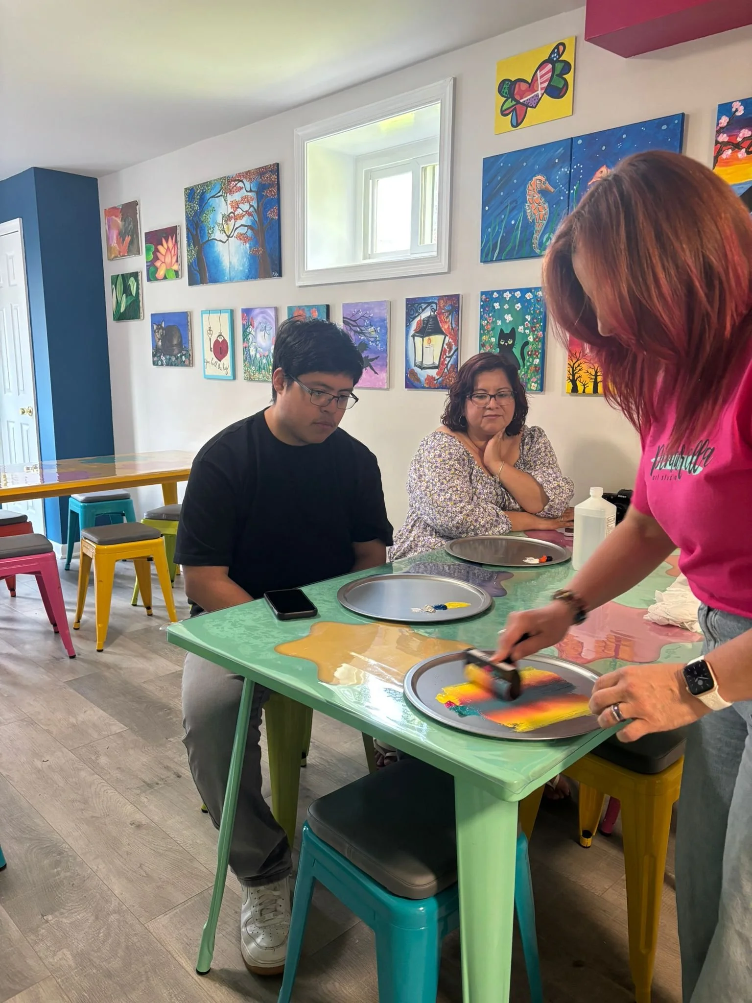 People preparing or serving colorful rainbow-designed food or art project at a green table in a brightly decorated room with paintings on the wall.