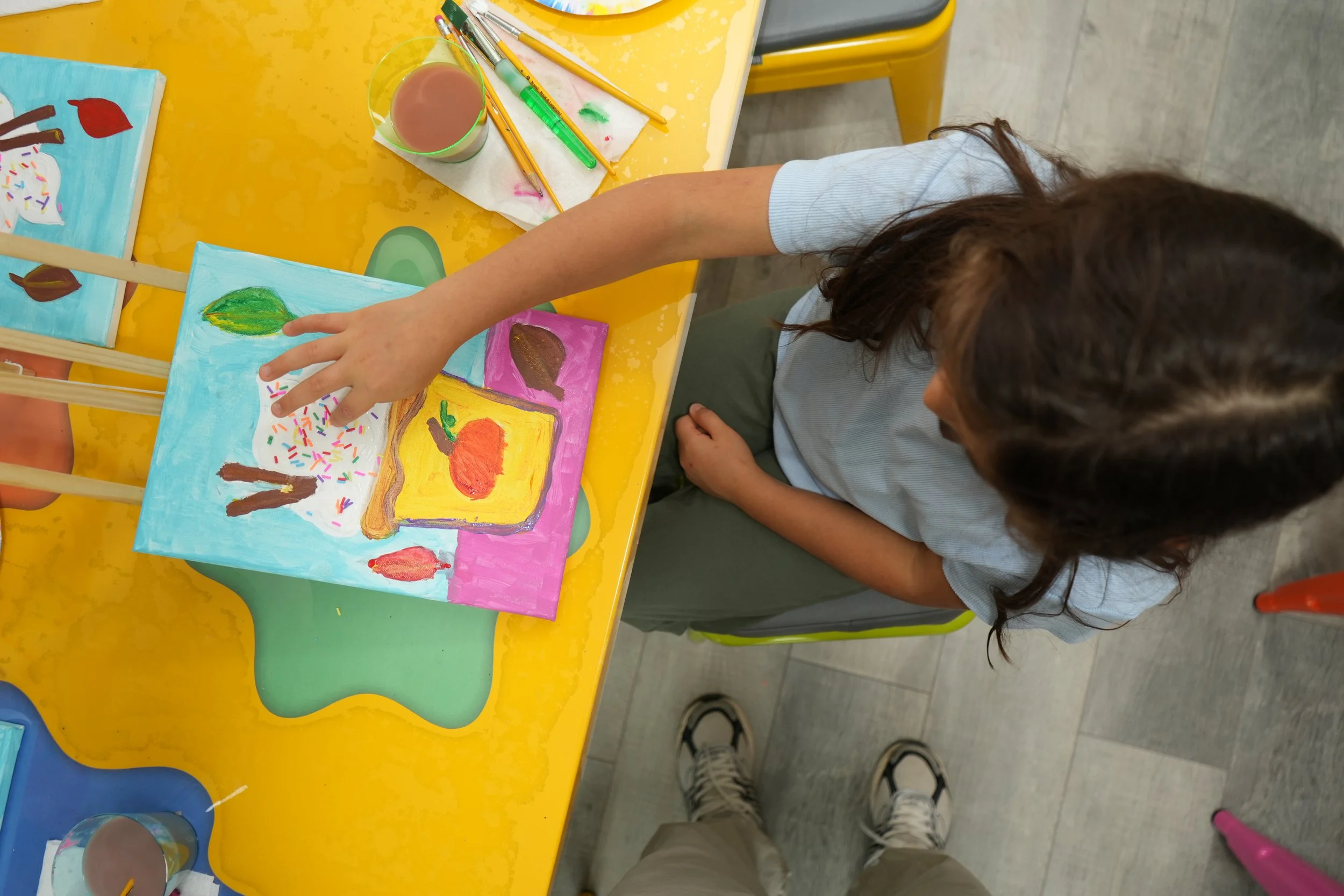 A young girl painting on colorful canvases at a yellow table, with brushes, a cup of water, and paint supplies nearby.
