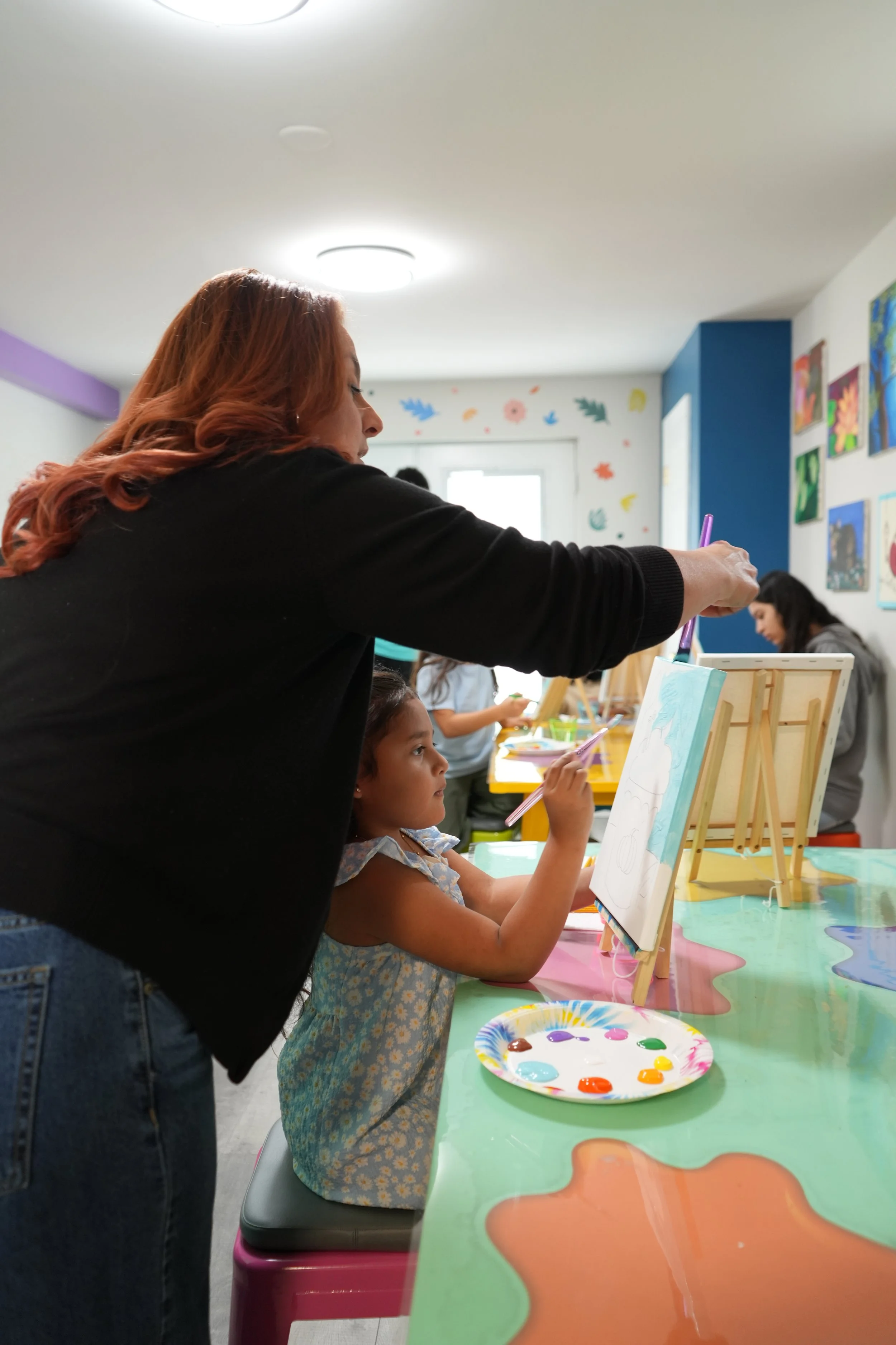 A woman with red hair assisting a young girl with dark hair, painting on a canvas at an art class. Other children are visible in the background, with paintings hanging on the wall and art supplies on the table.