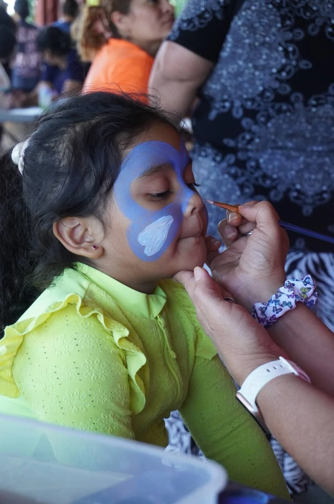 A young girl with face paint being painted by an artist at a festive outdoor event.