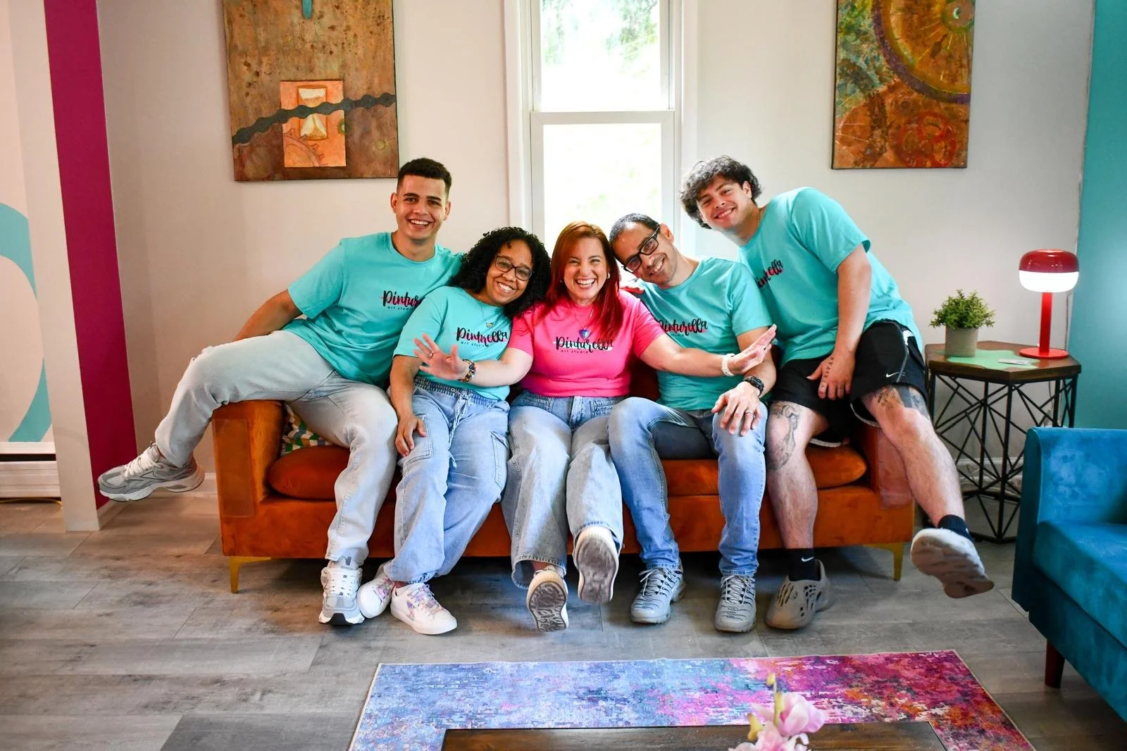 A group of six diverse people sitting on a sofa in a brightly decorated living room, smiling and posing for a photo.
