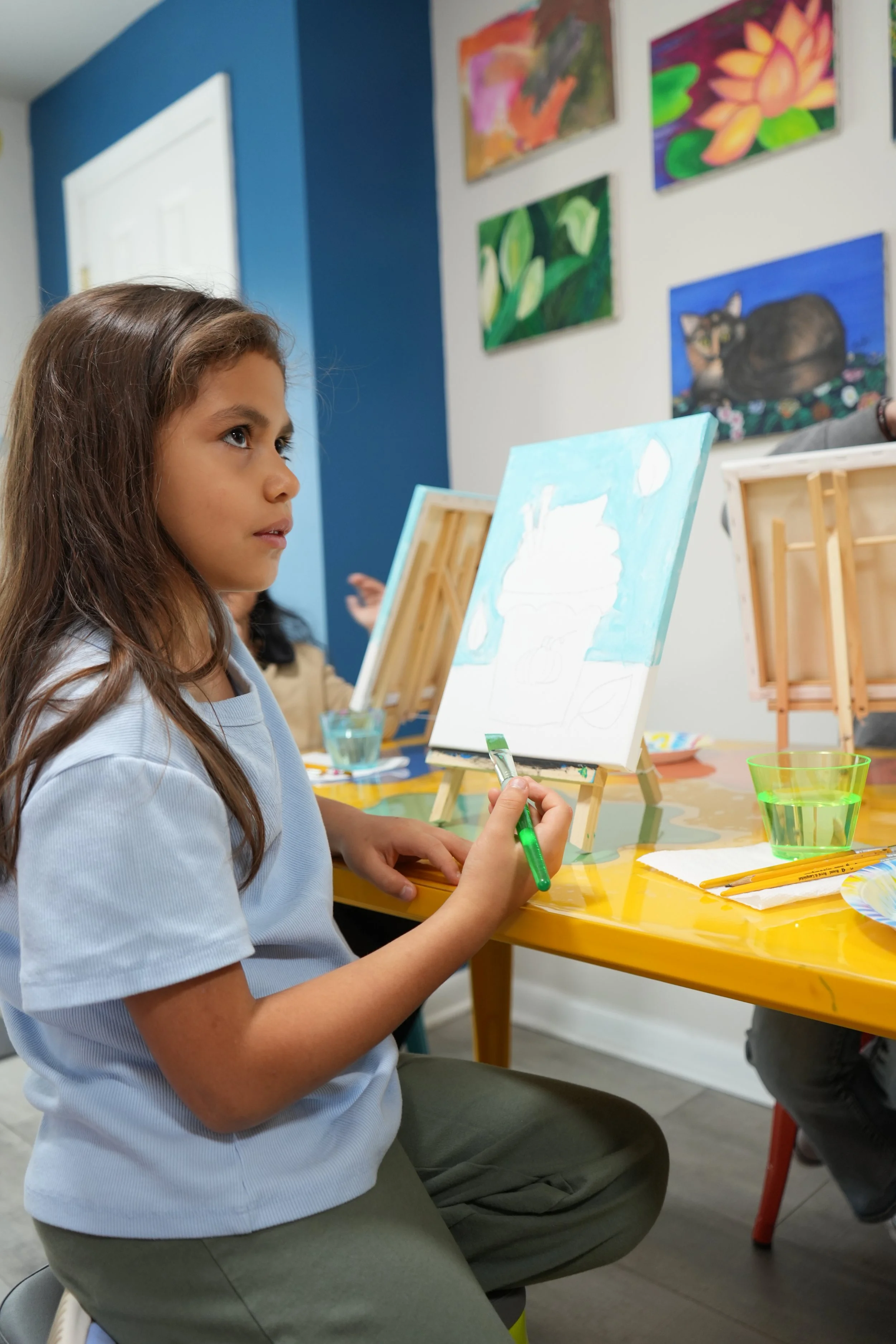 A young girl with long brown hair sitting at a yellow table, painting on a canvas, with several paintings of flowers and a cat hanging on the wall behind her. She is holding a green paintbrush and wearing a light blue shirt.