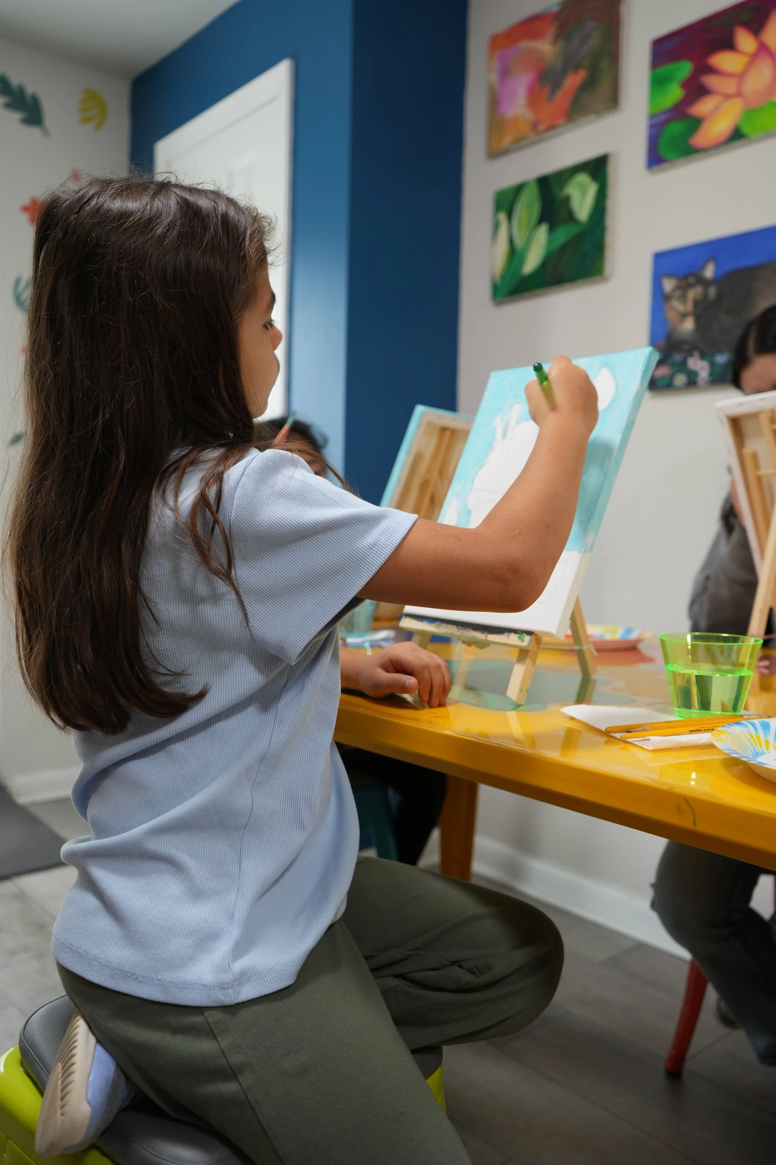 A young girl with long brown hair wearing a light blue shirt and gray pants, sitting on a gray stool, painting on a canvas set on an easel. She is holding a paintbrush and focused on her artwork. The background shows colorful paintings on the wall an